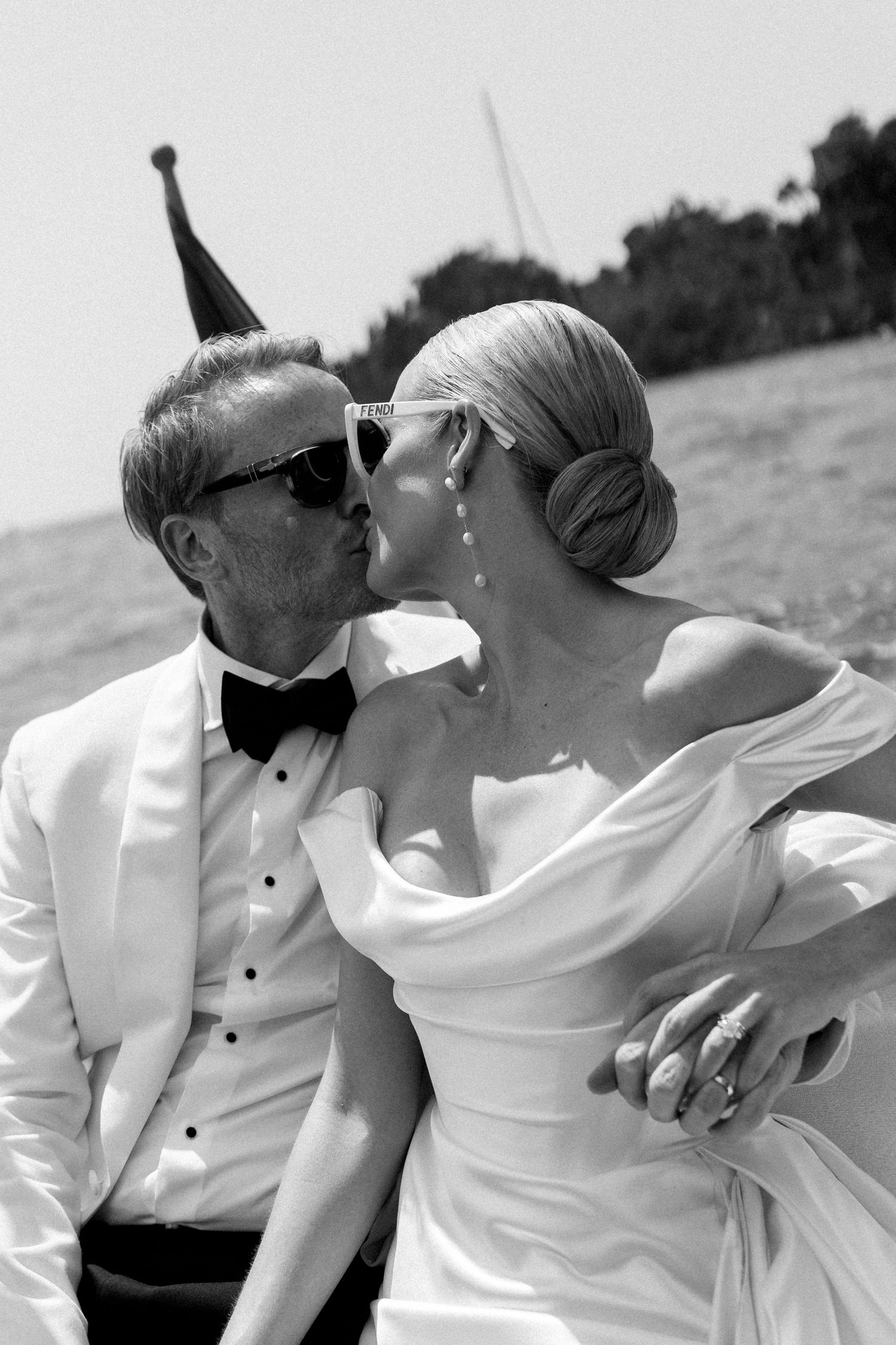 Bride and groom in sunglasses share a kiss while seated outdoors on their wedding day.