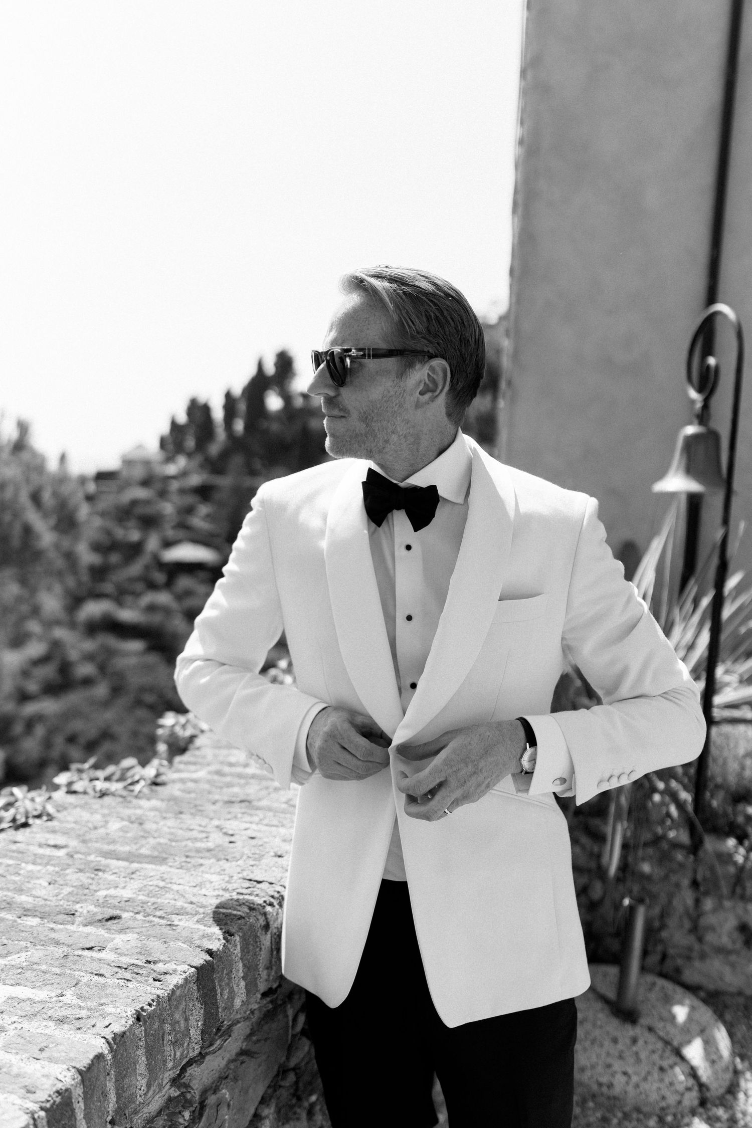 Groom in white tuxedo jacket and sunglasses adjusting jacket outdoors in black and white photo.