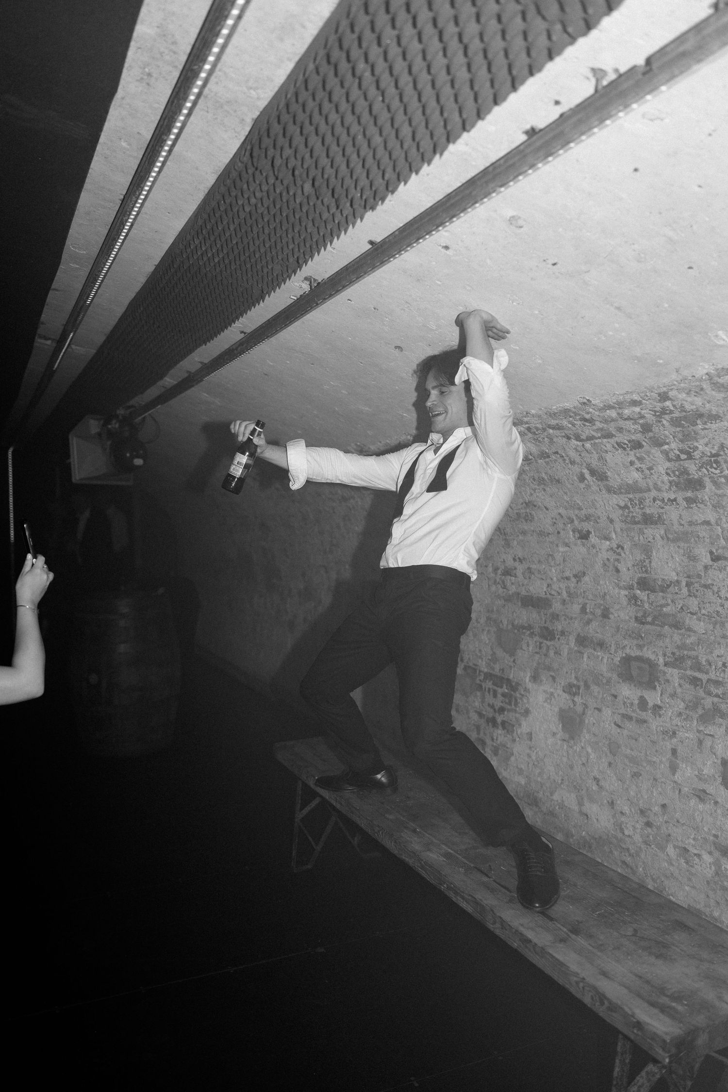 Black and white photo of a man in suspenders dancing energetically on a wooden bench in a dimly lit venue.