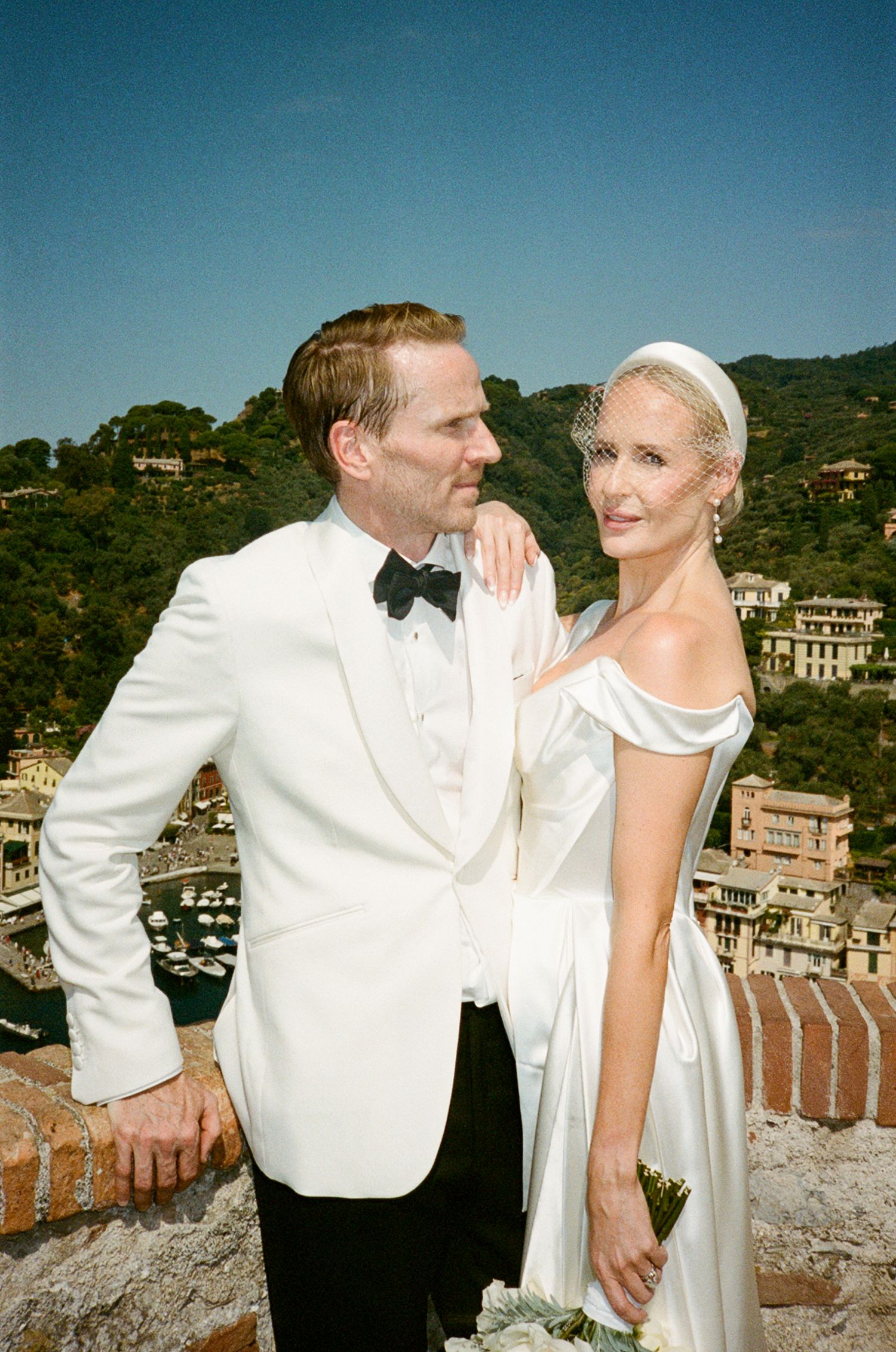 Bride and groom in white wedding attire pose with Italian coastal village backdrop.