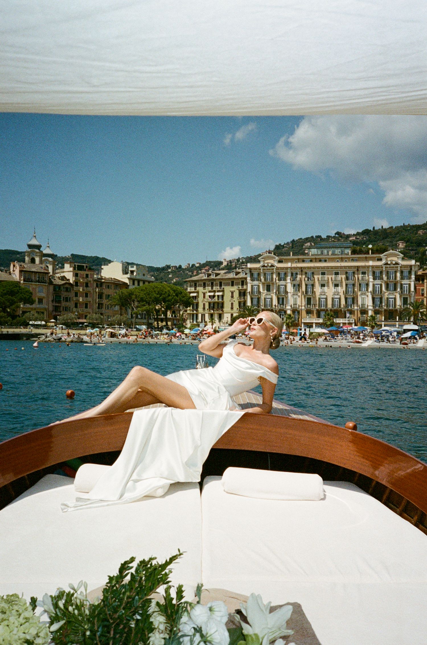 Bride in white gown reclines on luxury wooden boat bow with scenic Italian Riviera coastline behind.