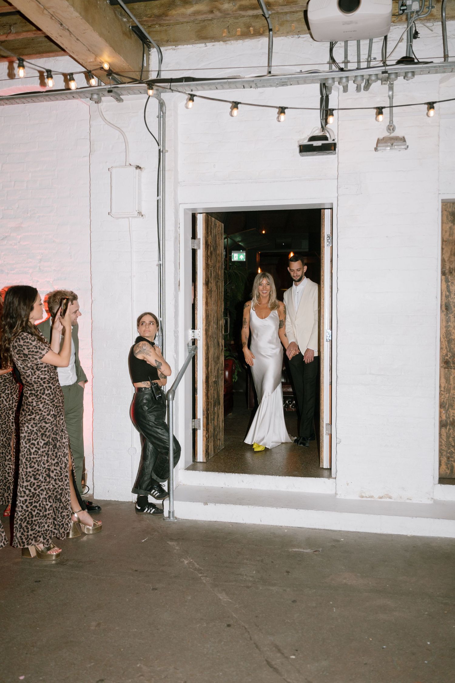 Wedding guests and a bride in white mingle in an industrial-style venue with string lights overhead.