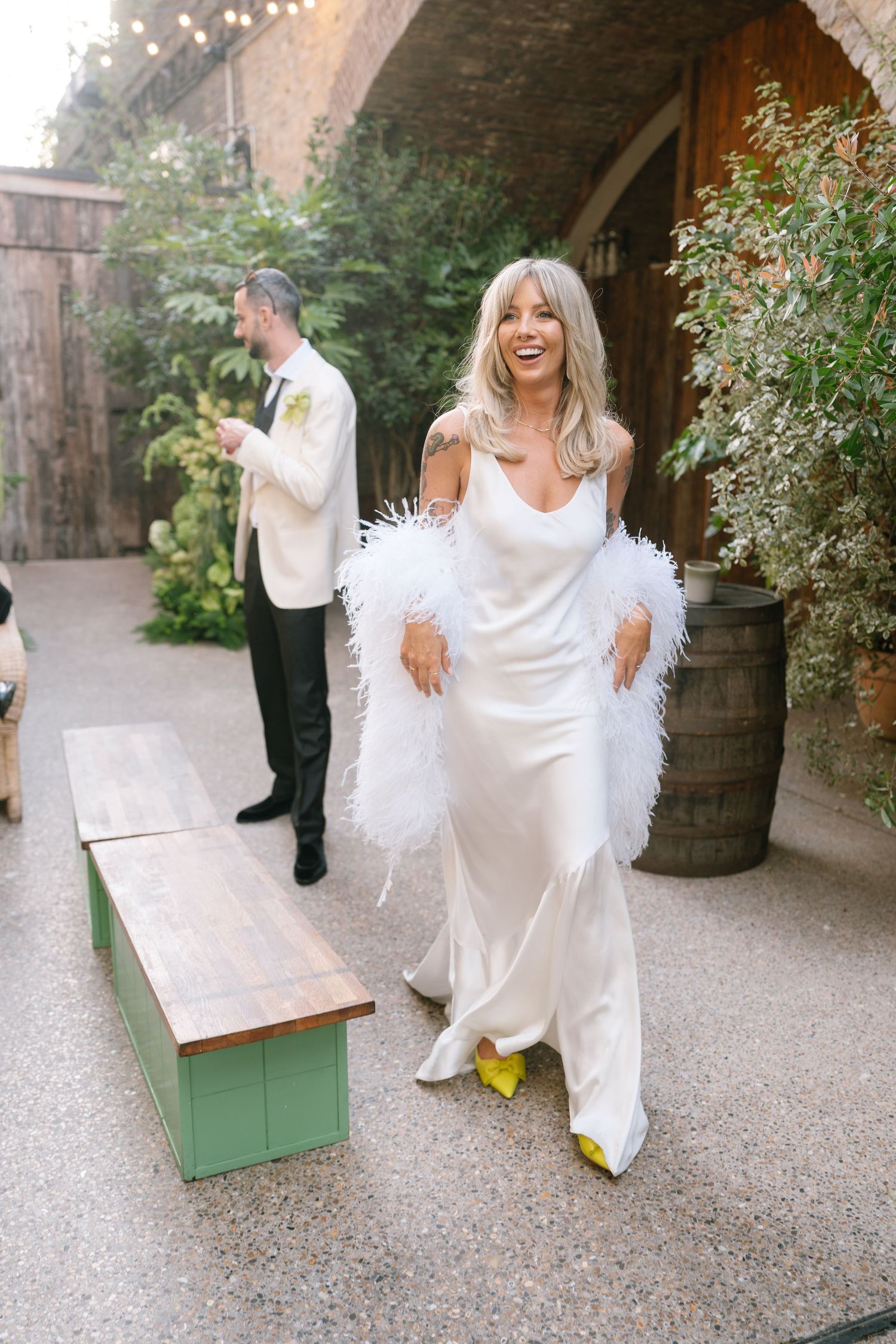 A bride in a white silk dress and feathered shawl poses playfully near wine barrels at an outdoor venue.