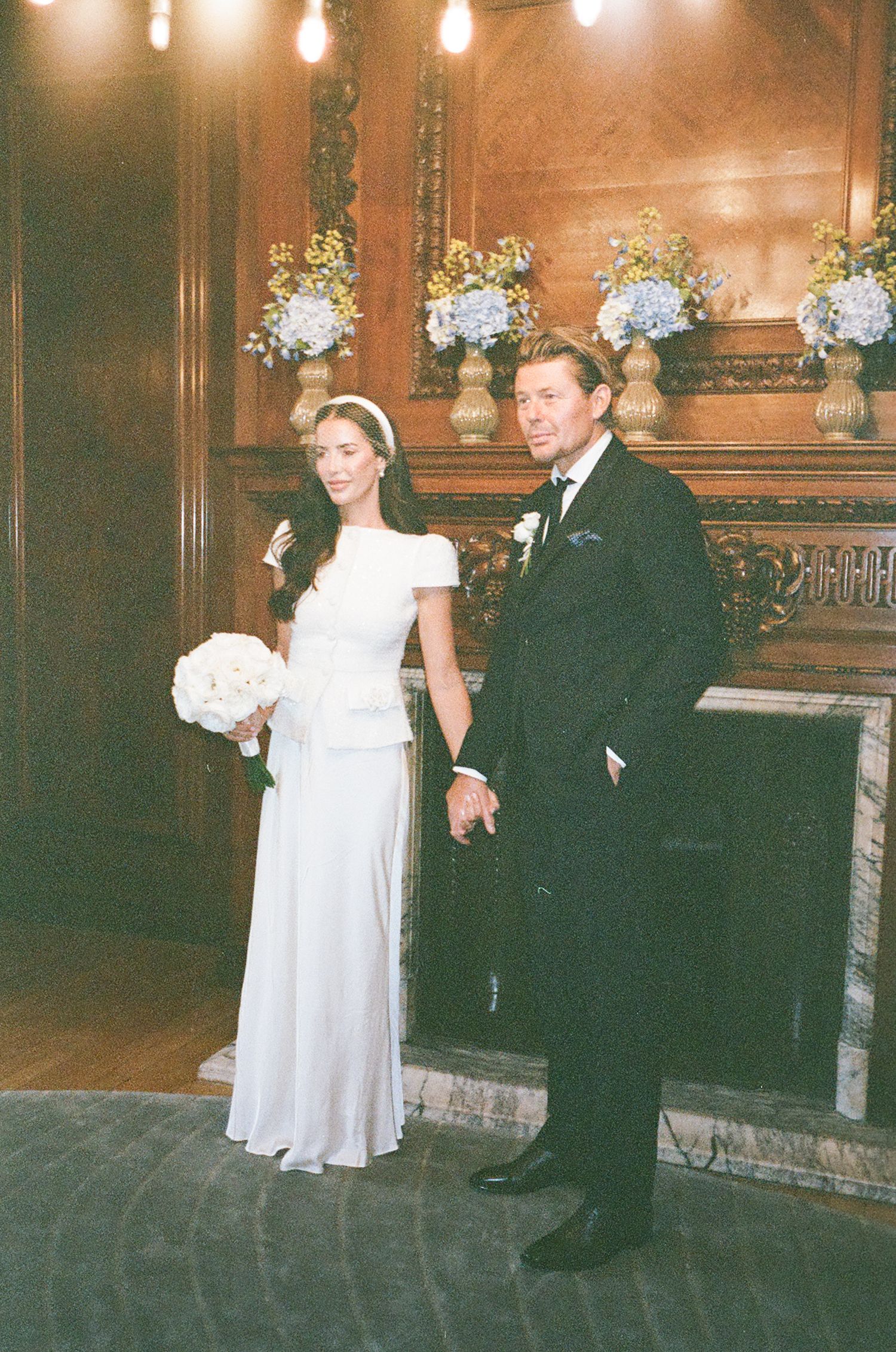 A wedding couple poses in an elegant venue with ornate wood paneling and floral arrangements illuminated by warm lighting.