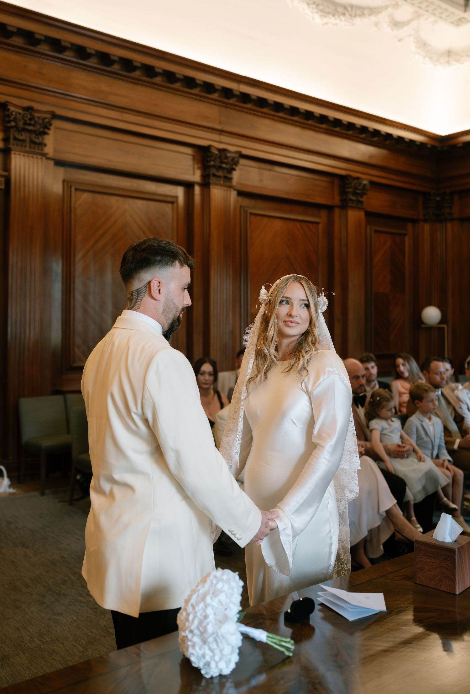 A wedding ceremony takes place in an elegant wood-paneled room with ornate Corinthian columns.