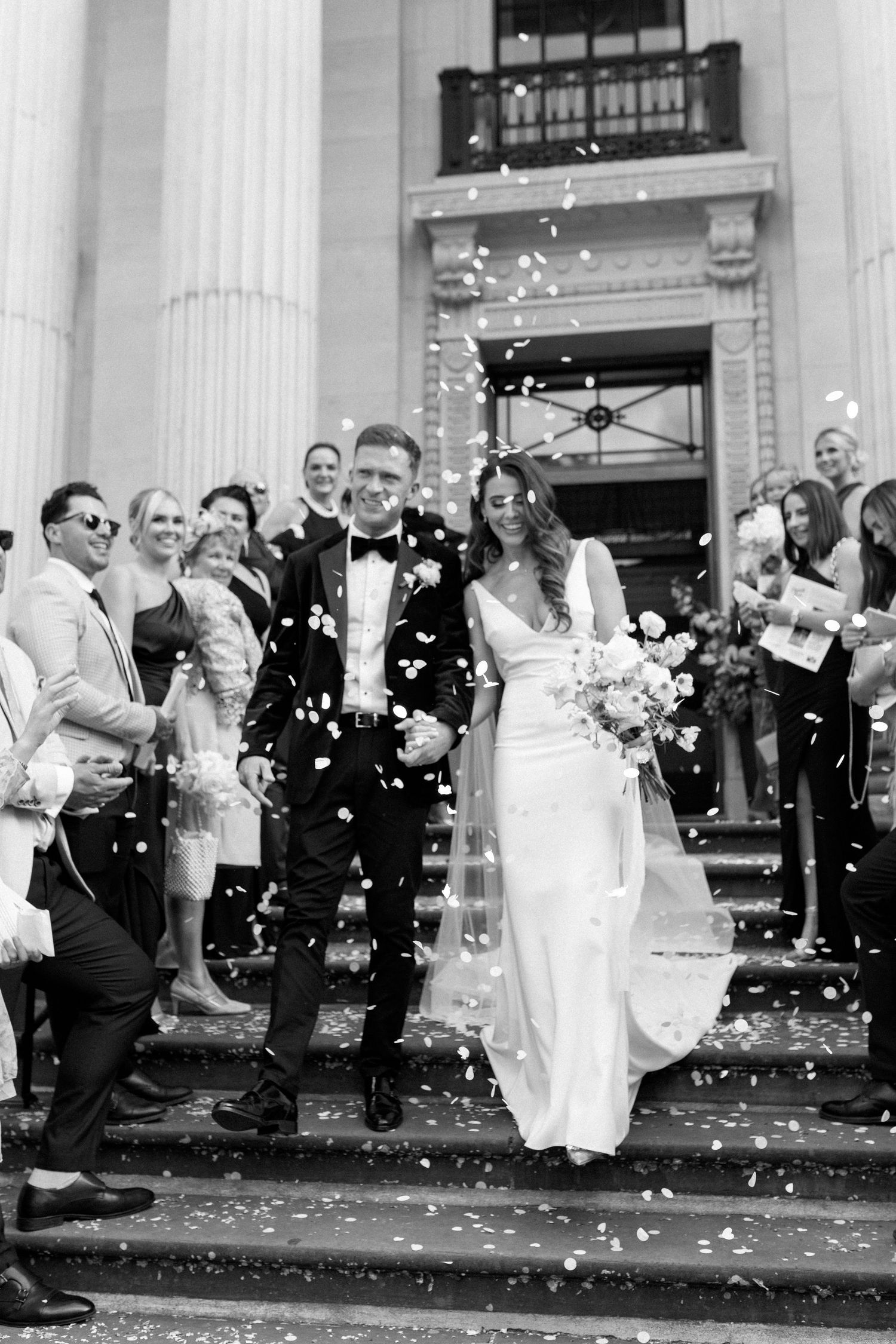 Wedding celebration with flower petals being tossed outside classical building entrance.