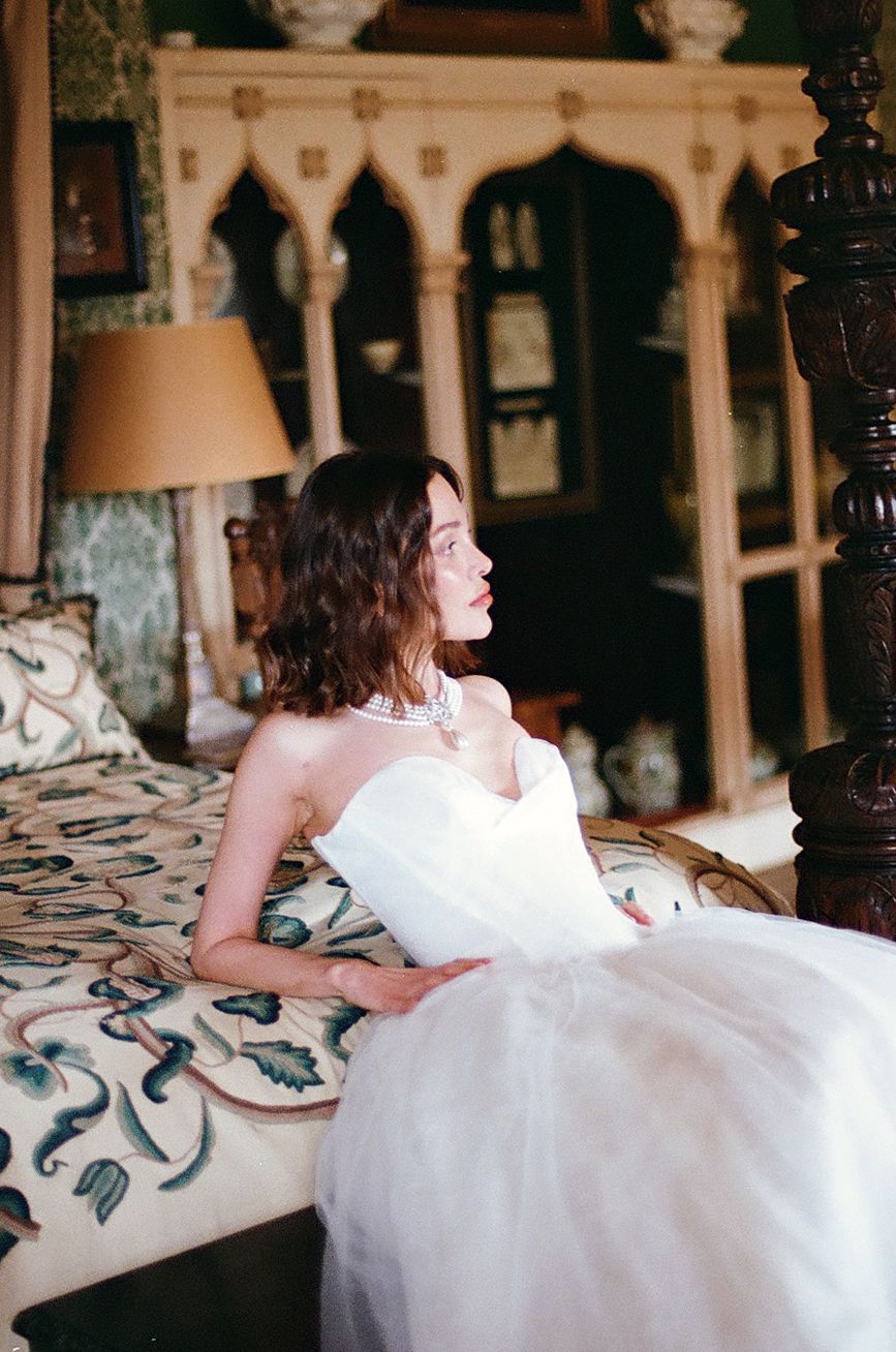 A person in a white dress reclines on a floral patterned bed in an ornate vintage-style room.