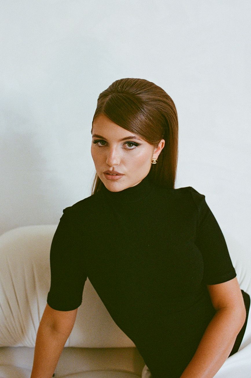 Woman in black turtleneck top with sleek brown hair poses elegantly against a white background.
