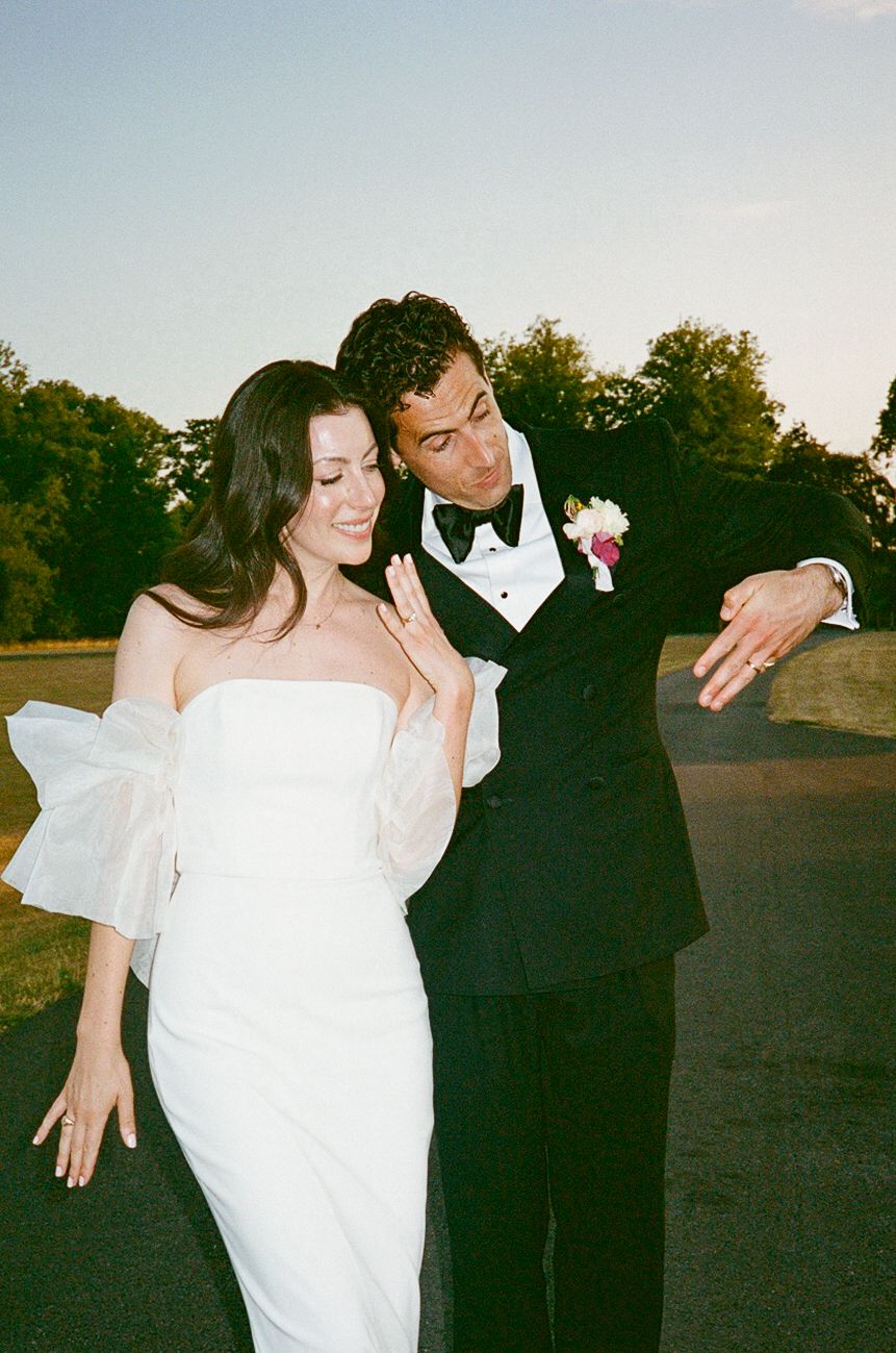 Bride in white strapless gown with bow sleeves and groom in black tuxedo showing off wedding rings outdoors.