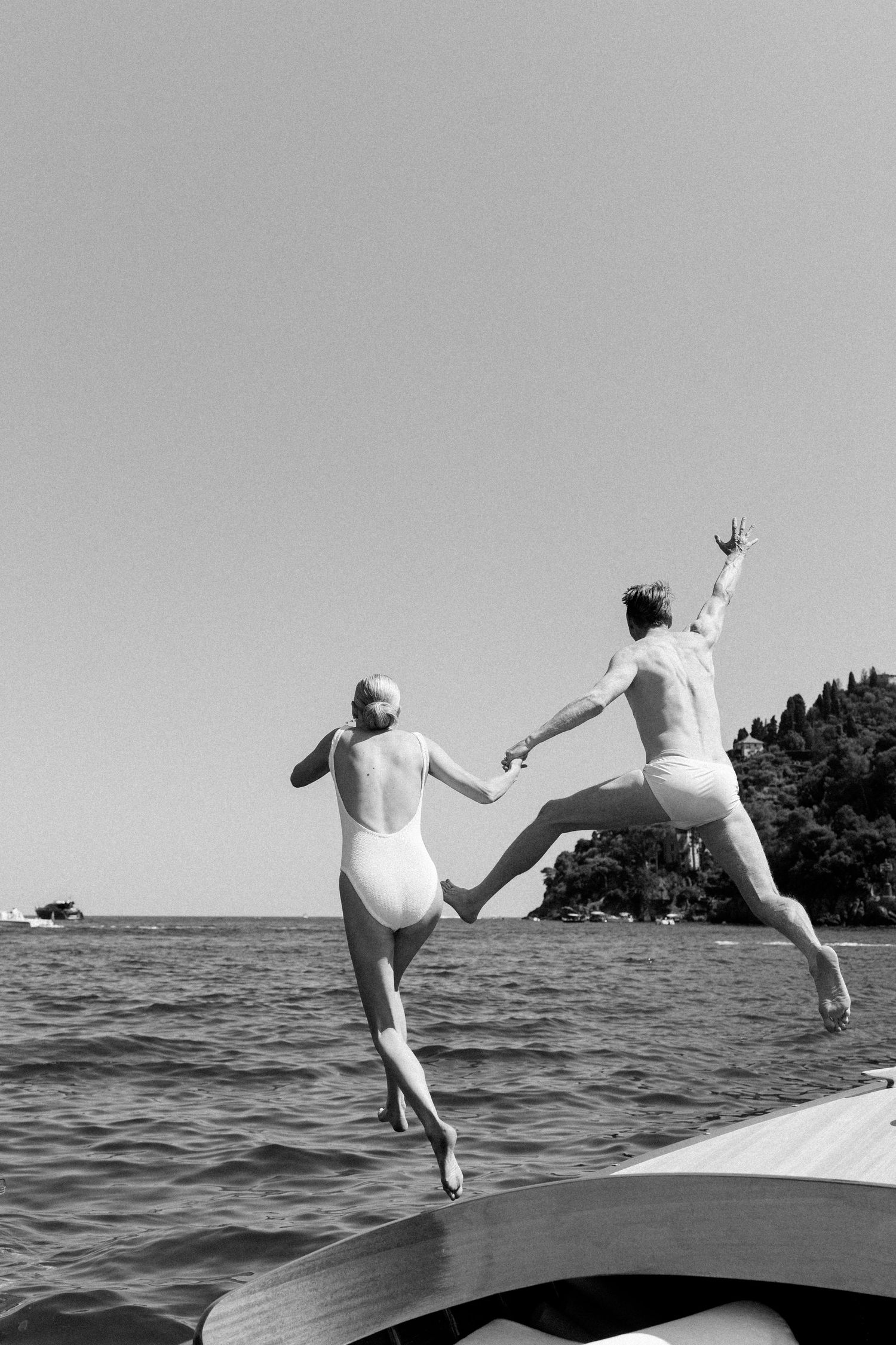 Black and white photo of a couple holding hands jumping off a boat into the sea on a sunny day.