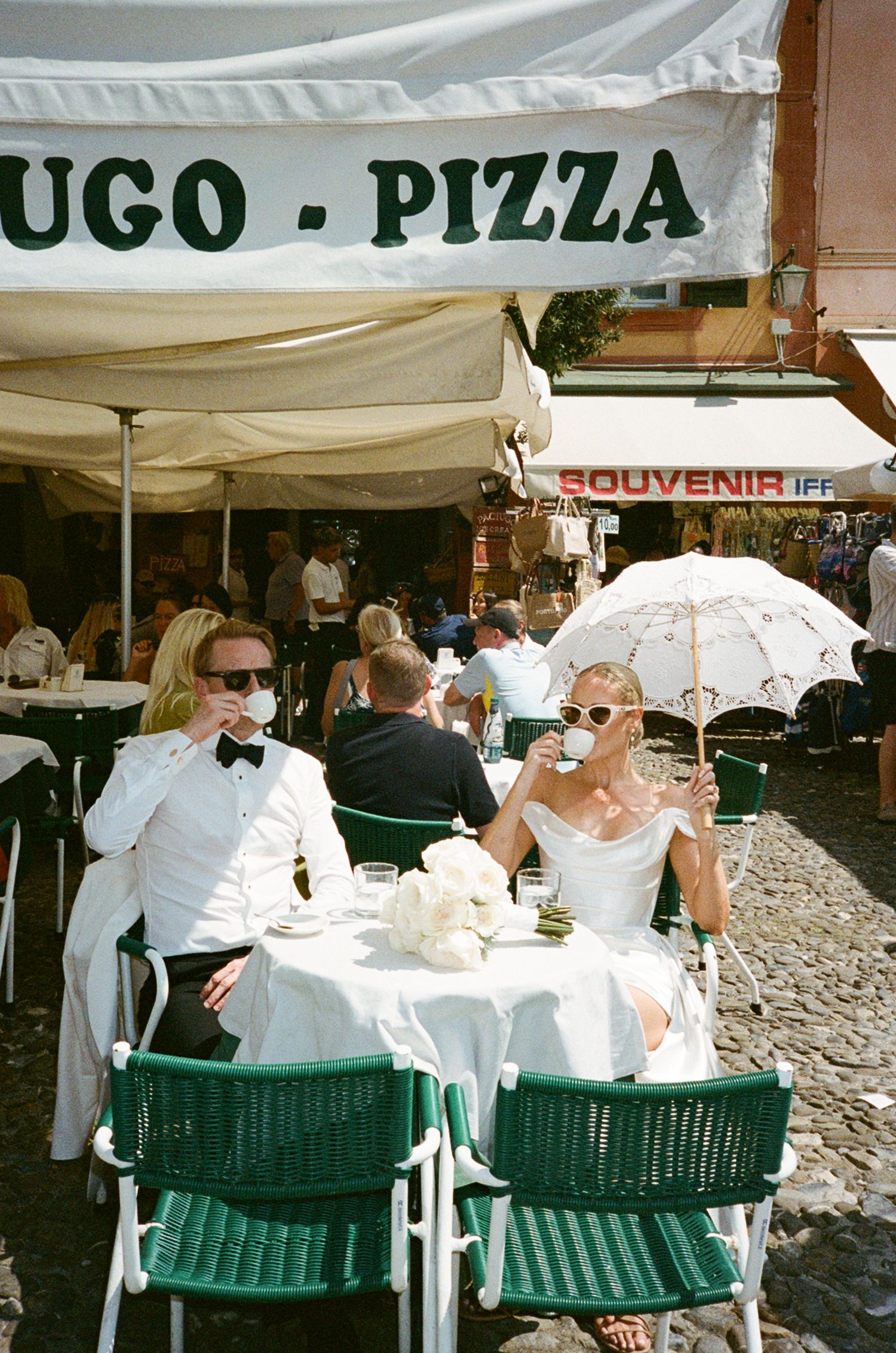 Elegant couple in white attire sipping coffee at outdoor pizza cafe table with green chairs and parasol.
