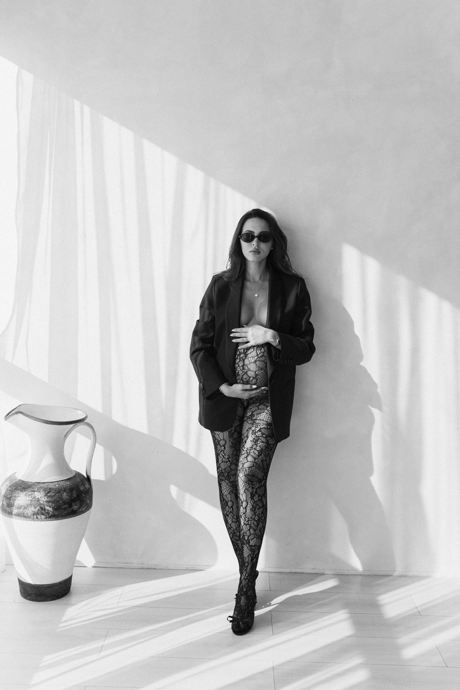 Pregnant woman in black blazer and lace tights poses against white wall with ceramic vase in black and white photo.
