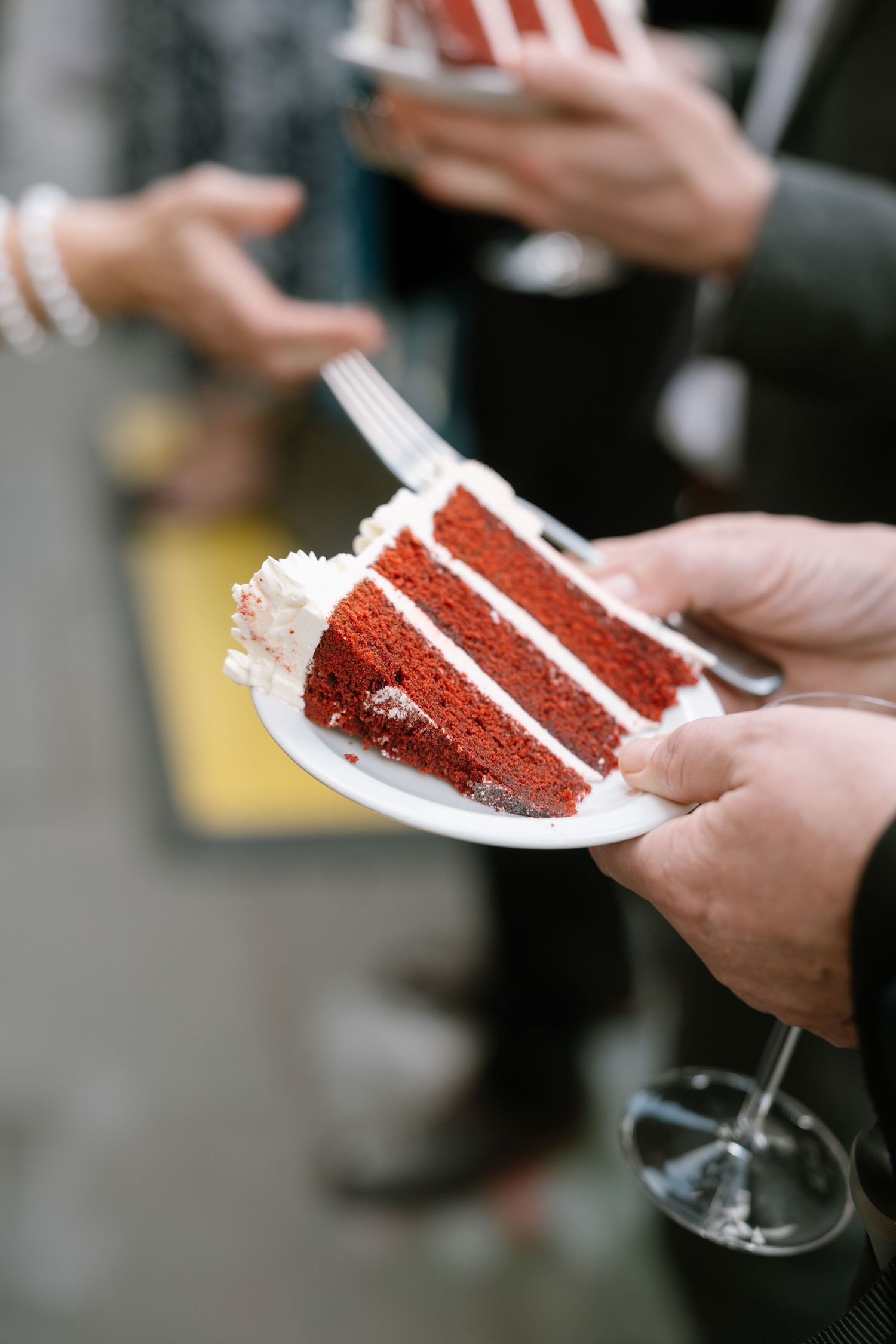 Multiple hands holding plates of red velvet cake slices at a wedding reception.