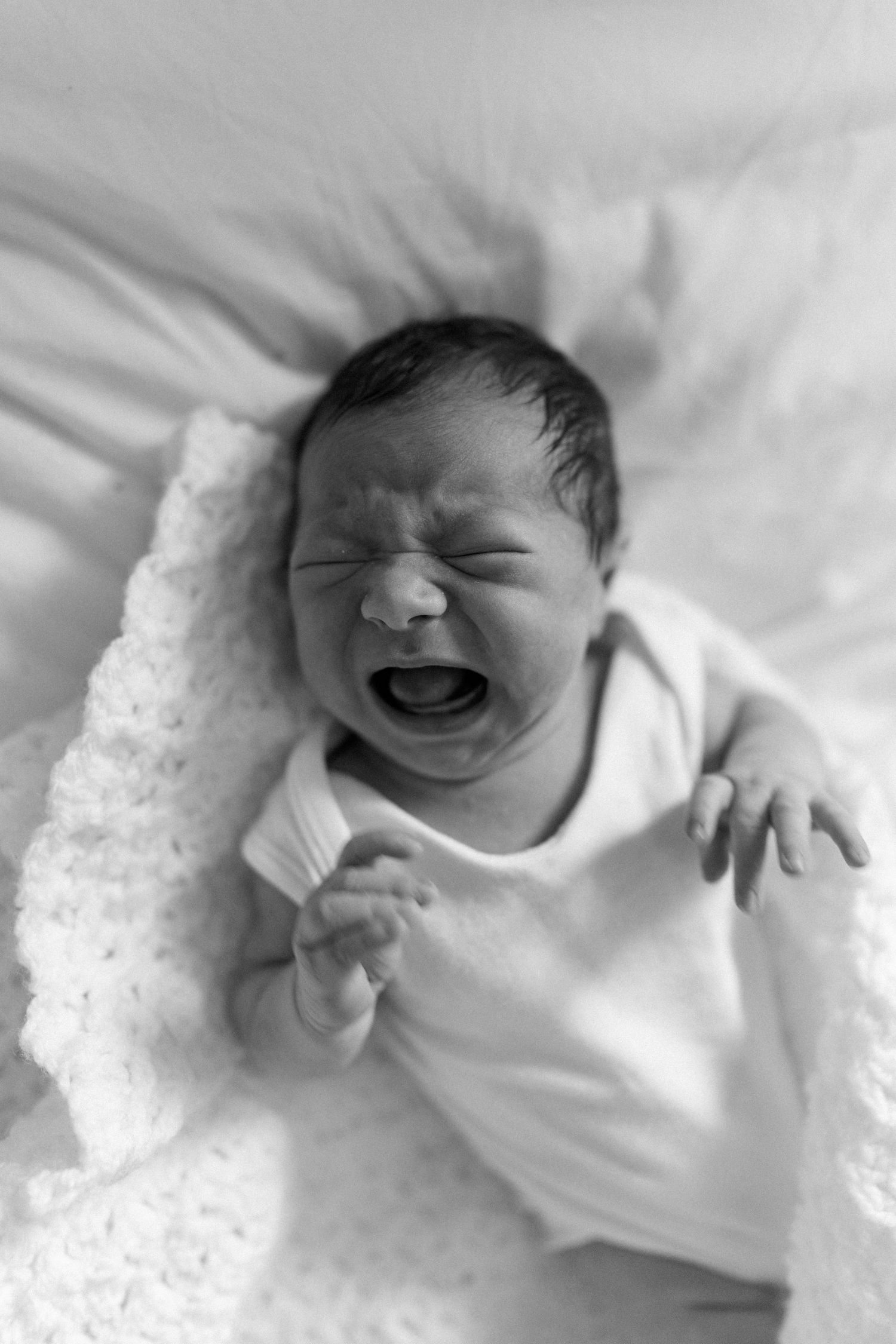 Laughing newborn baby in white onesie lying on white bedding.