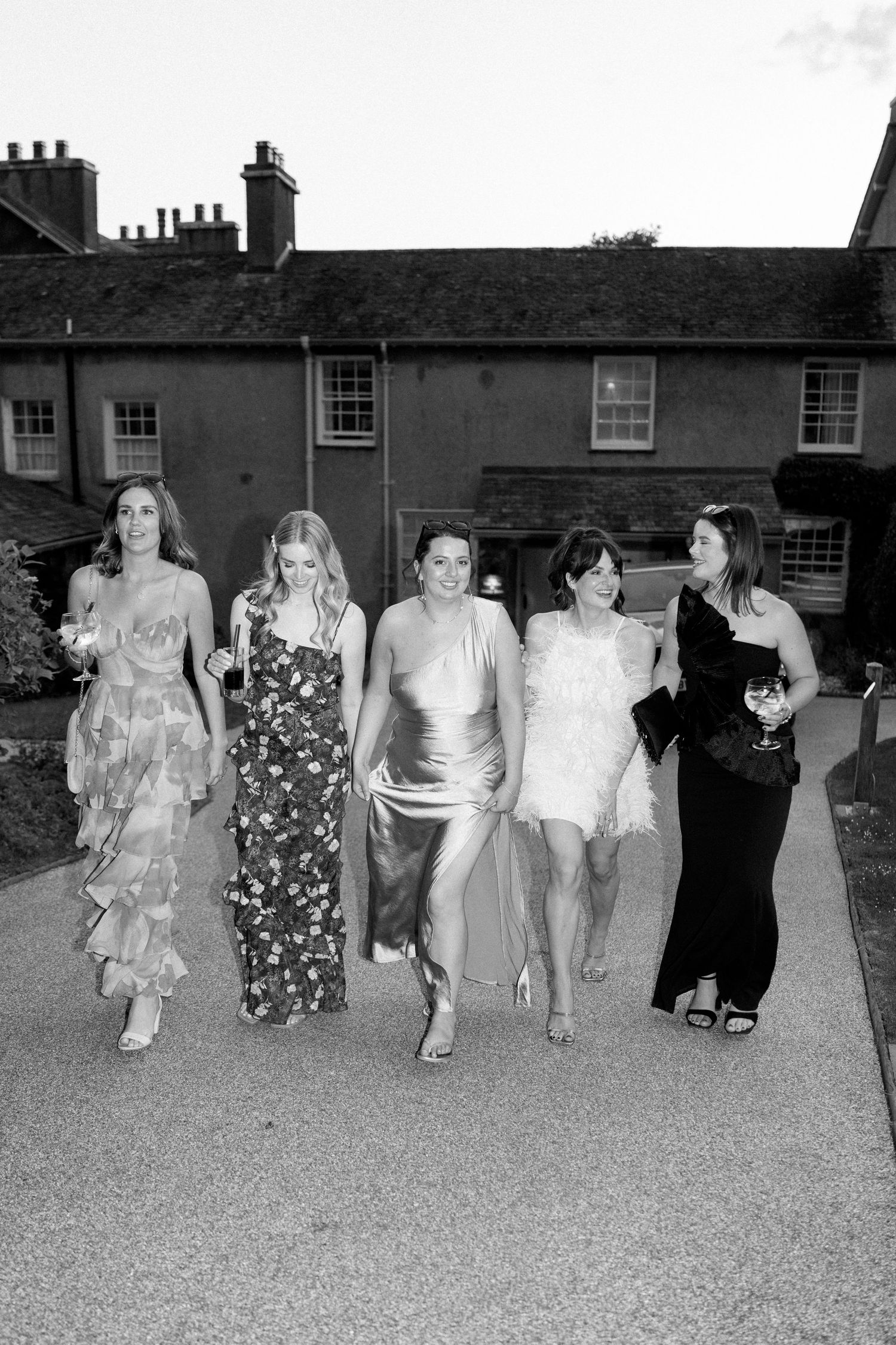 A group of friends in formal dresses walk together outside a traditional stone building.