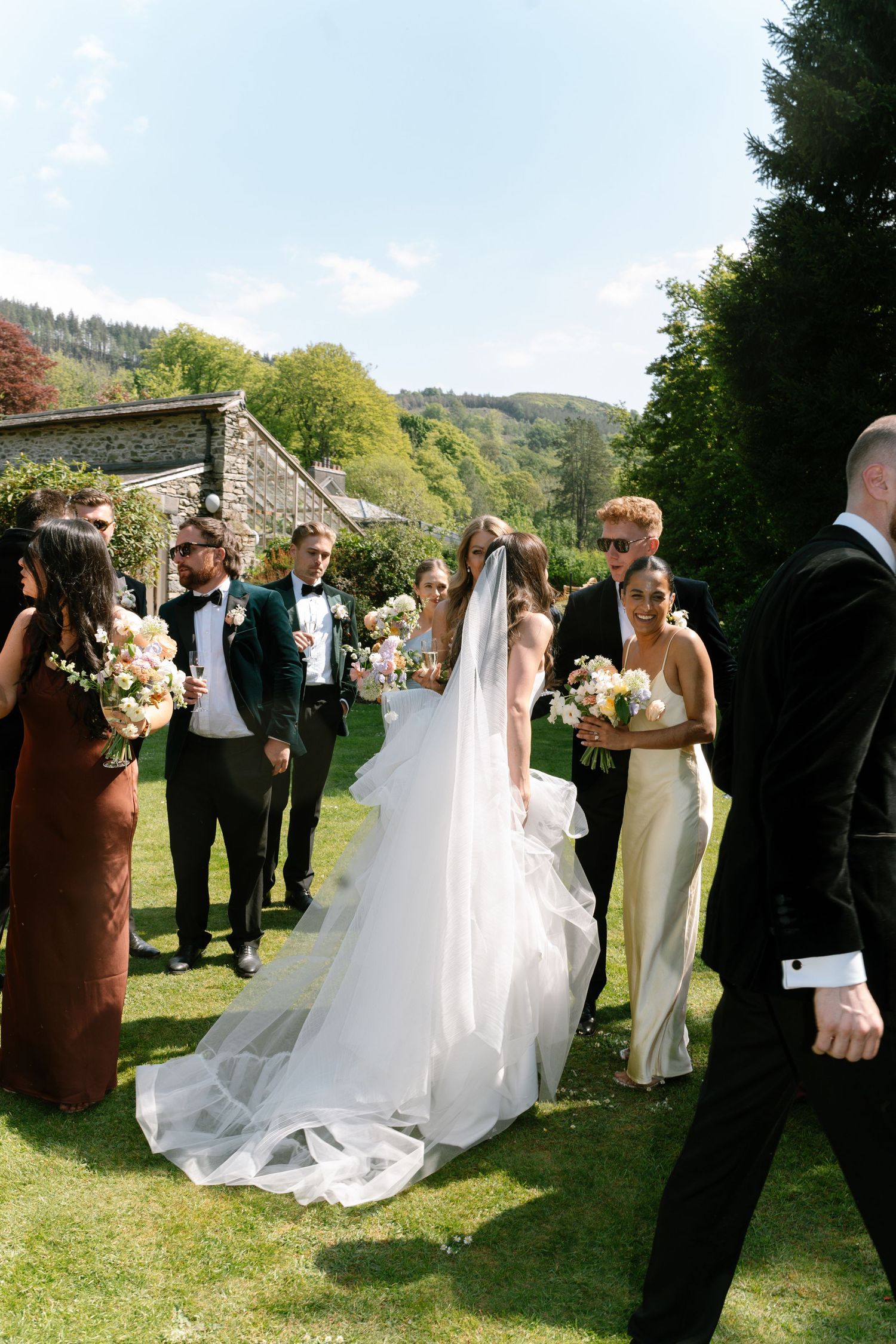 Elegant wedding party poses outdoors with stone building and mountain backdrop.