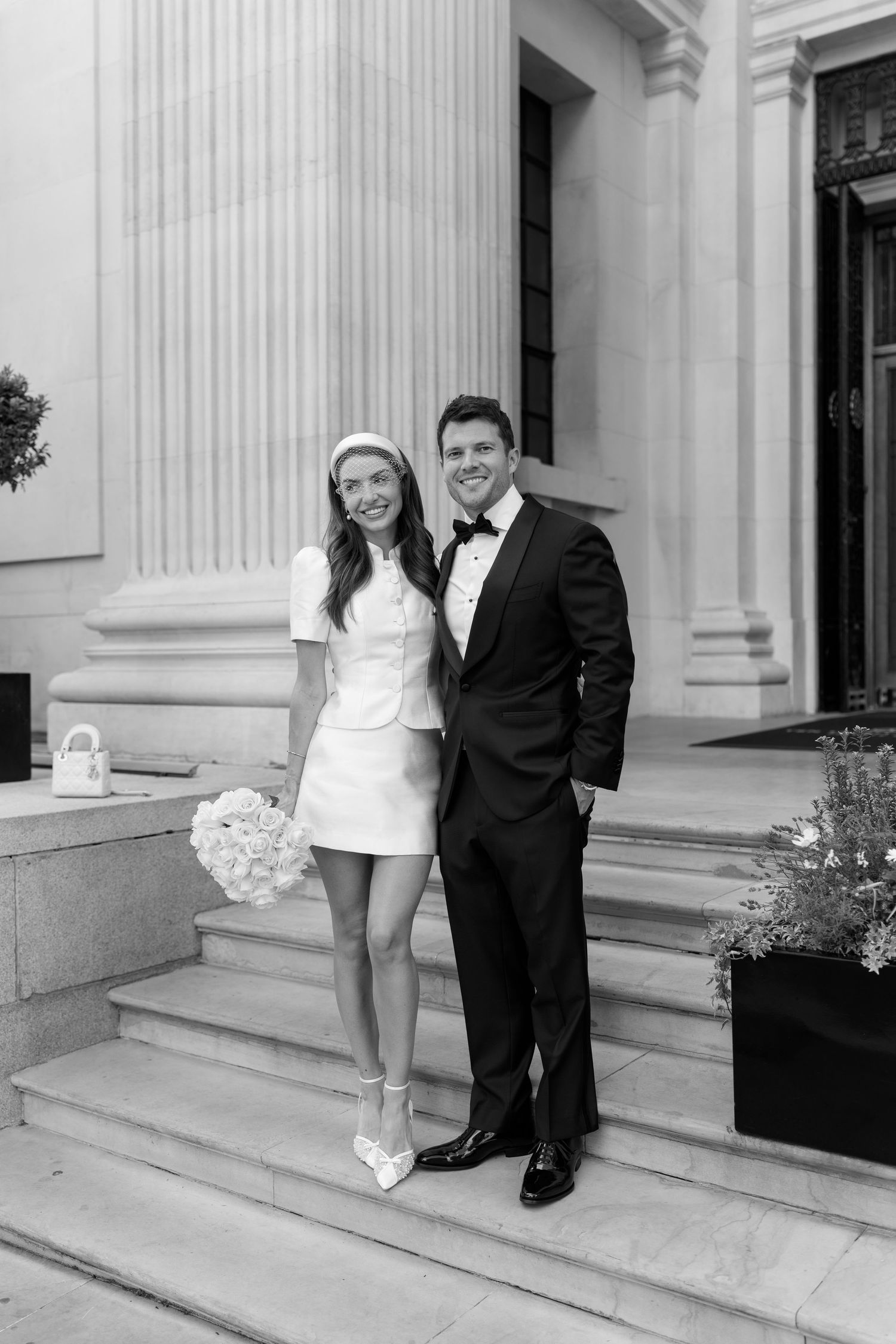 Bride in short white dress and groom in tuxedo posing on stone steps outside elegant building.