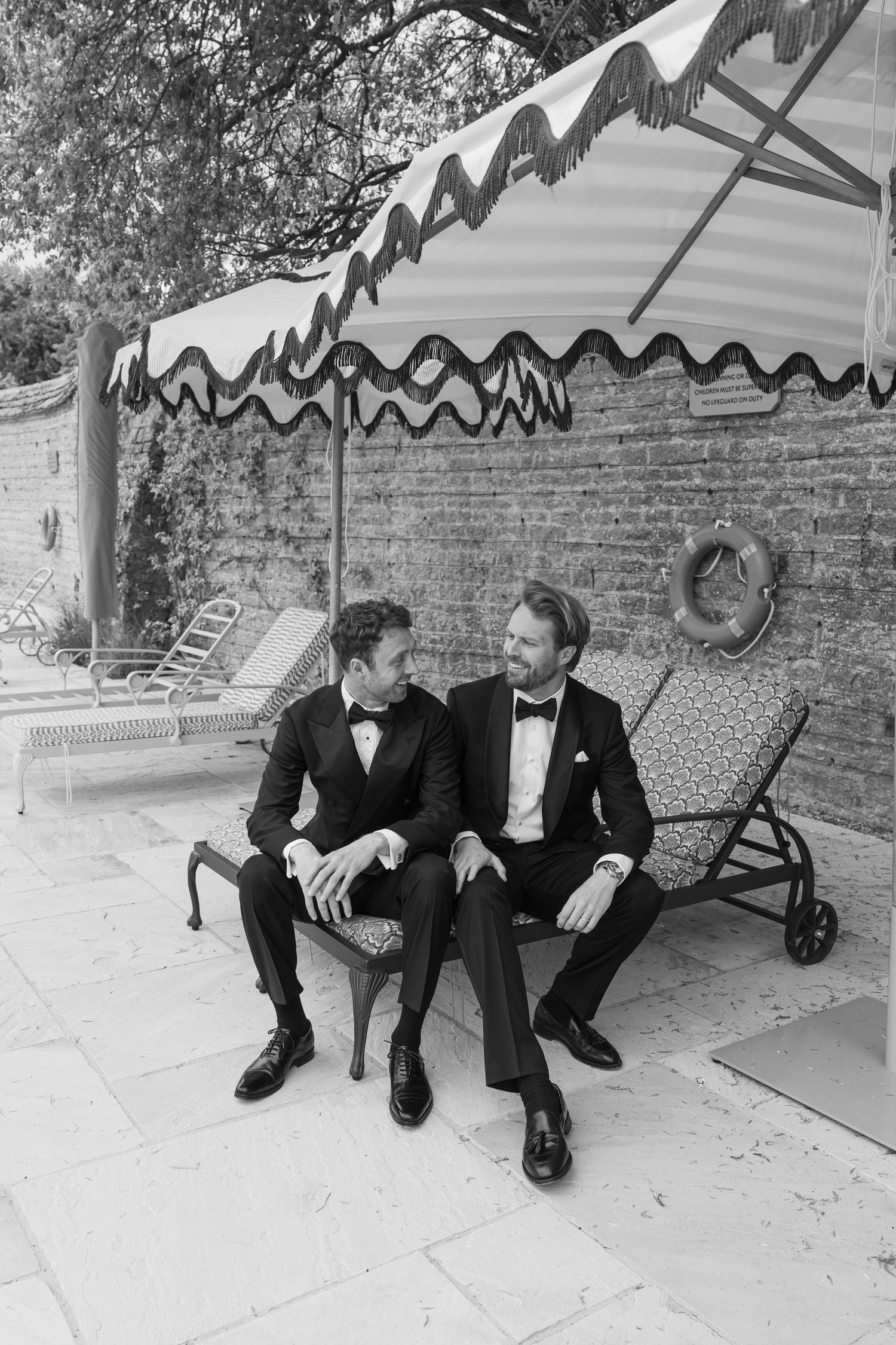 Two grooms in black tuxedos sharing an intimate moment seated under a striped poolside umbrella.