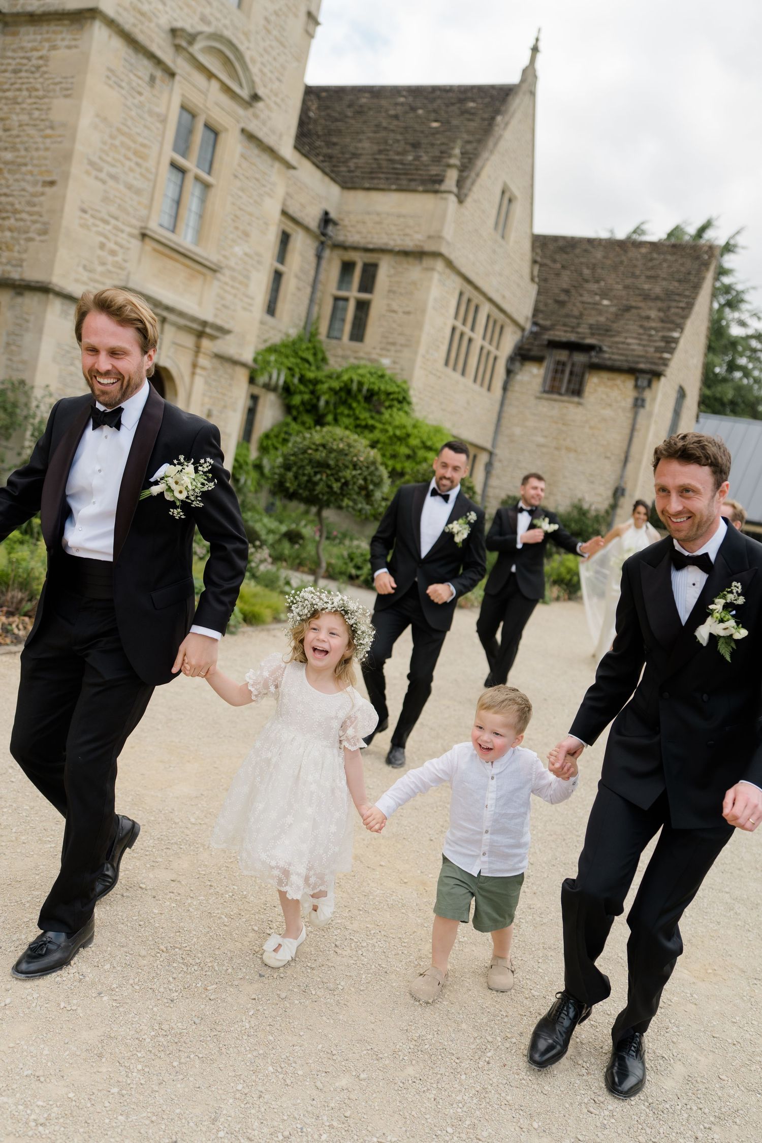 Groomsmen in black tuxedos walk with joyful flower girl and ring bearer at elegant stone manor wedding.