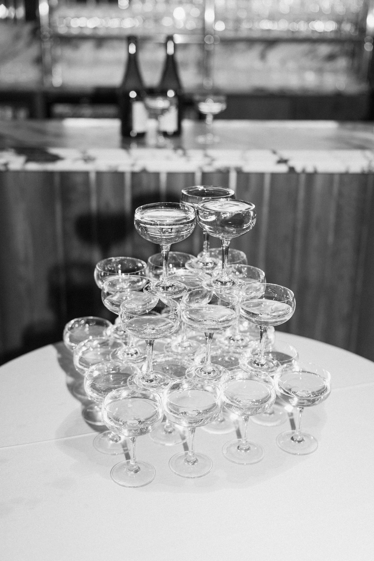 Black and white photo of elegant pyramid champagne tower with coupe glasses arranged on white tablecloth at wedding.