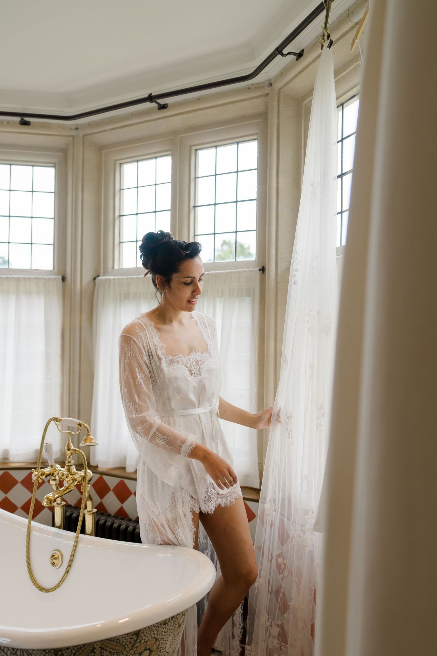 Bride in a white lace robe standing beside a vintage clawfoot bathtub near bay windows.