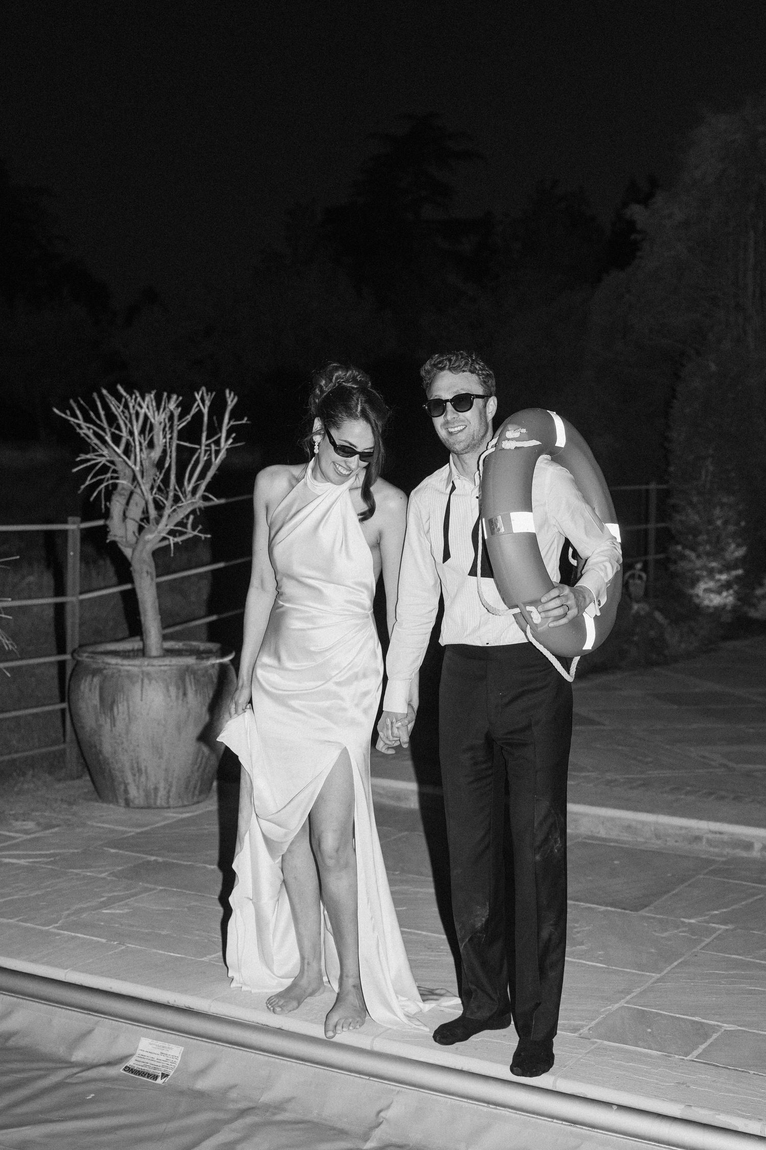 Bride and groom in sunglasses pose poolside at night in black and white holding life preserver ring.