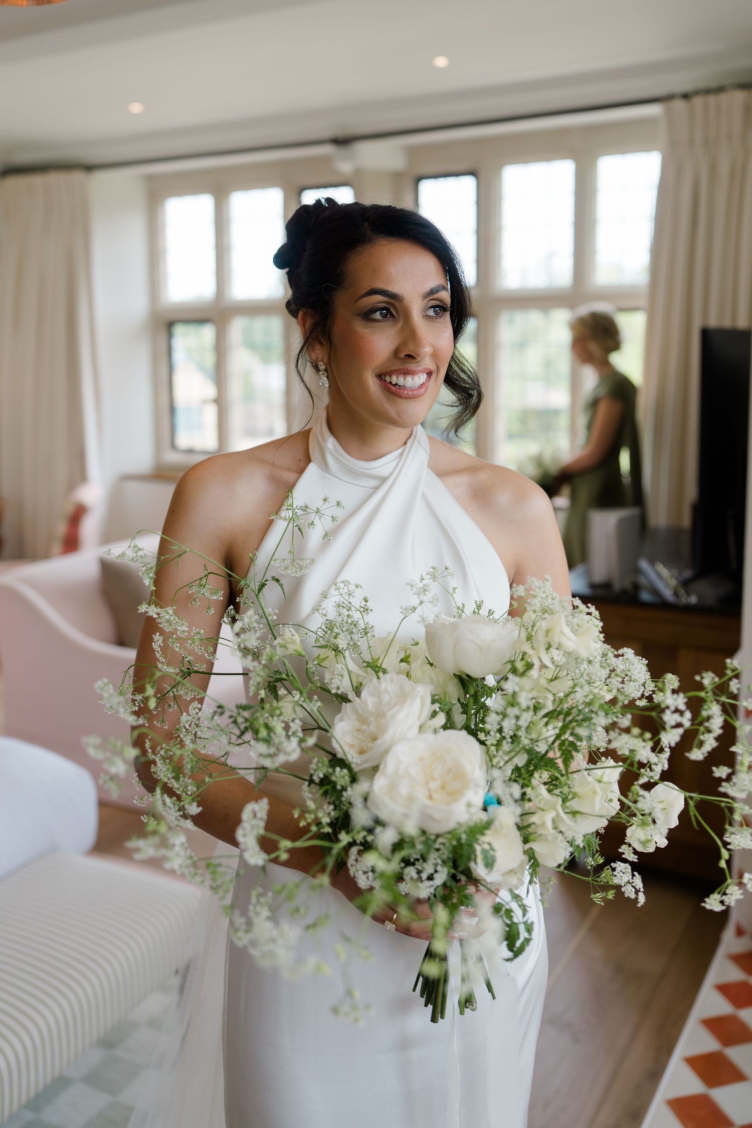 Smiling bride in halter neck wedding dress holds white floral bouquet indoors before ceremony.