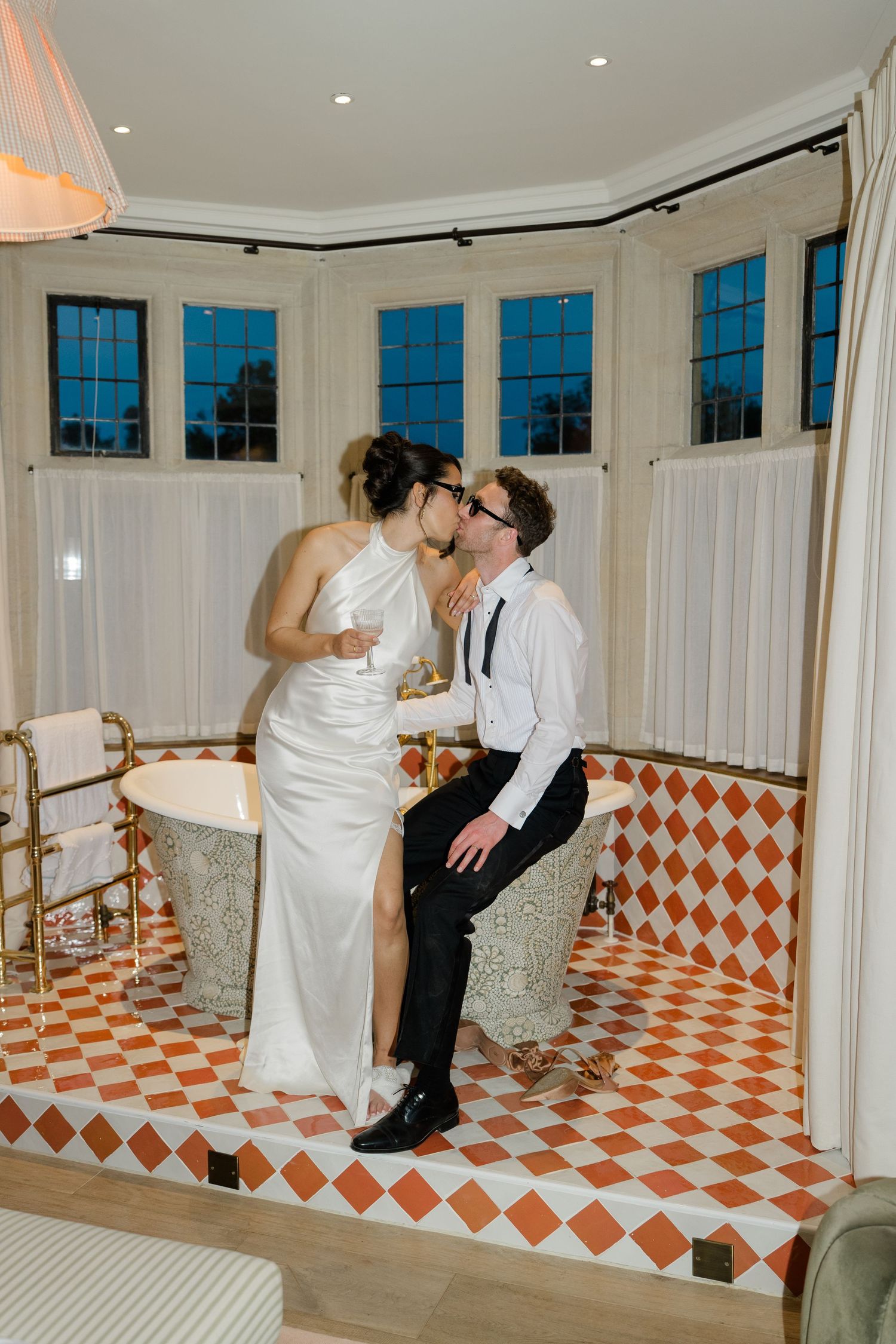 Bride and groom share a kiss while seated on a clawfoot tub in a stylish tiled bathroom.