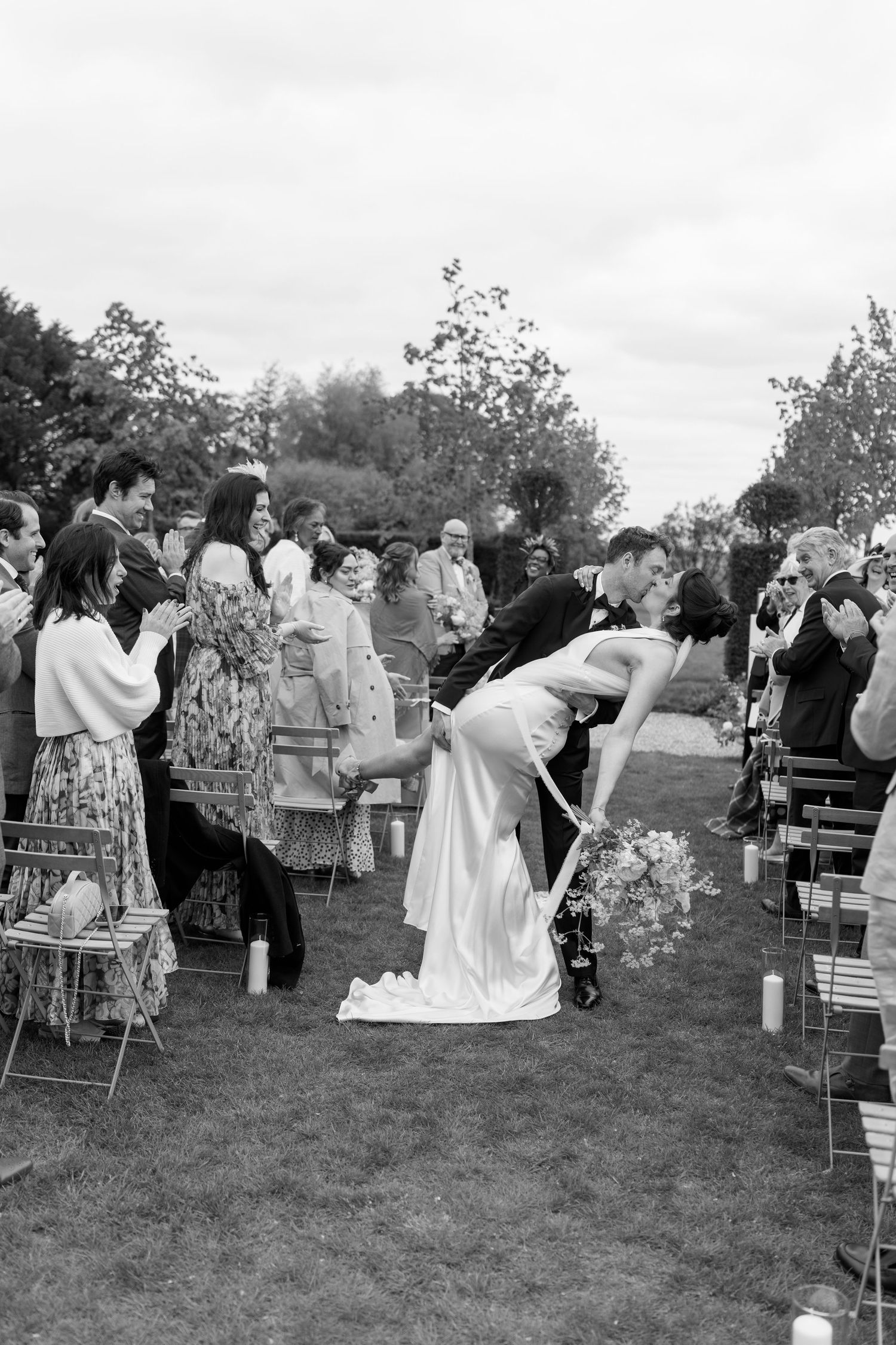 Groom dips bride in romantic kiss while guests cheer along the aisle after outdoor wedding ceremony.