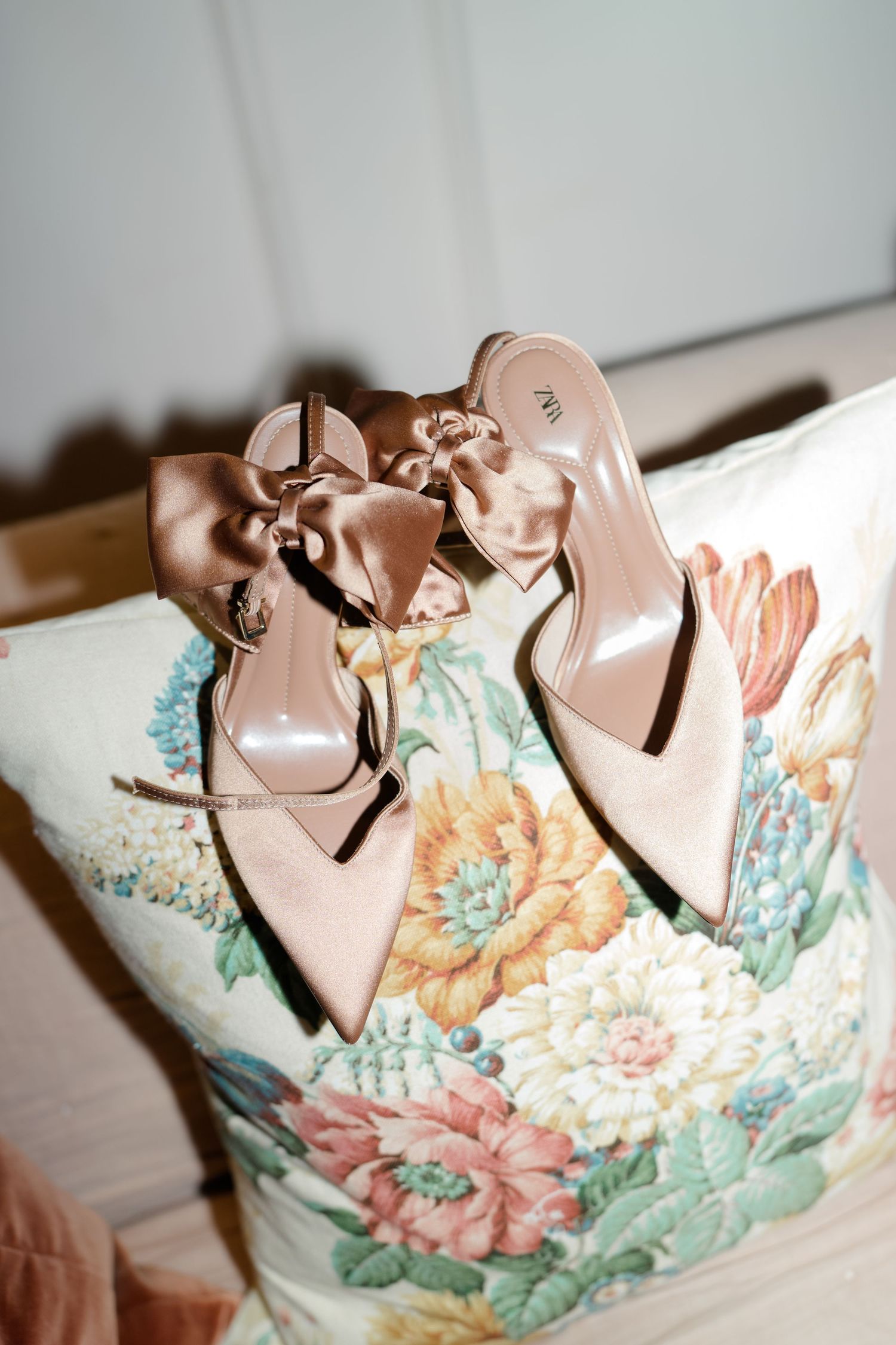 Blush pink satin heeled mules with bow detail resting on a floral embroidered cushion.