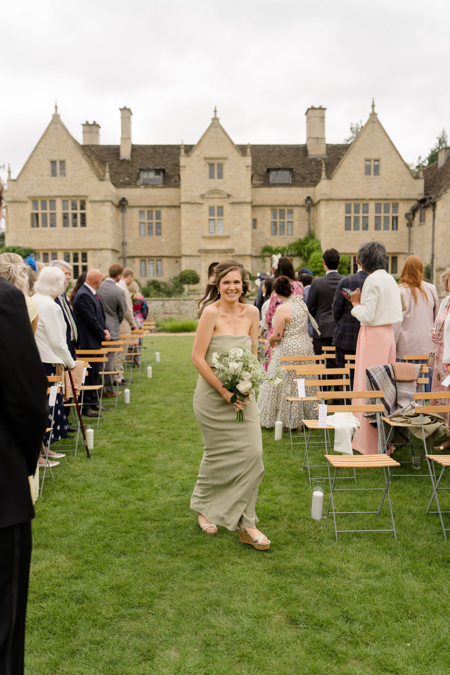 Bridesmaid in sage green dress walking down outdoor aisle at a Cotswolds manor wedding.
