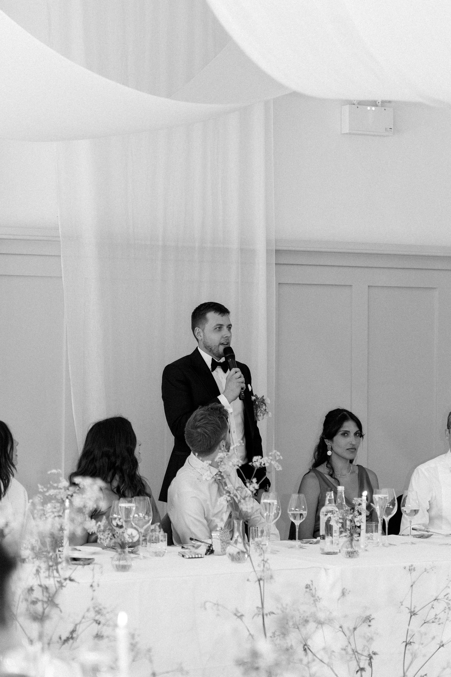 Groom delivering wedding speech to seated guests in elegant white draped reception venue.