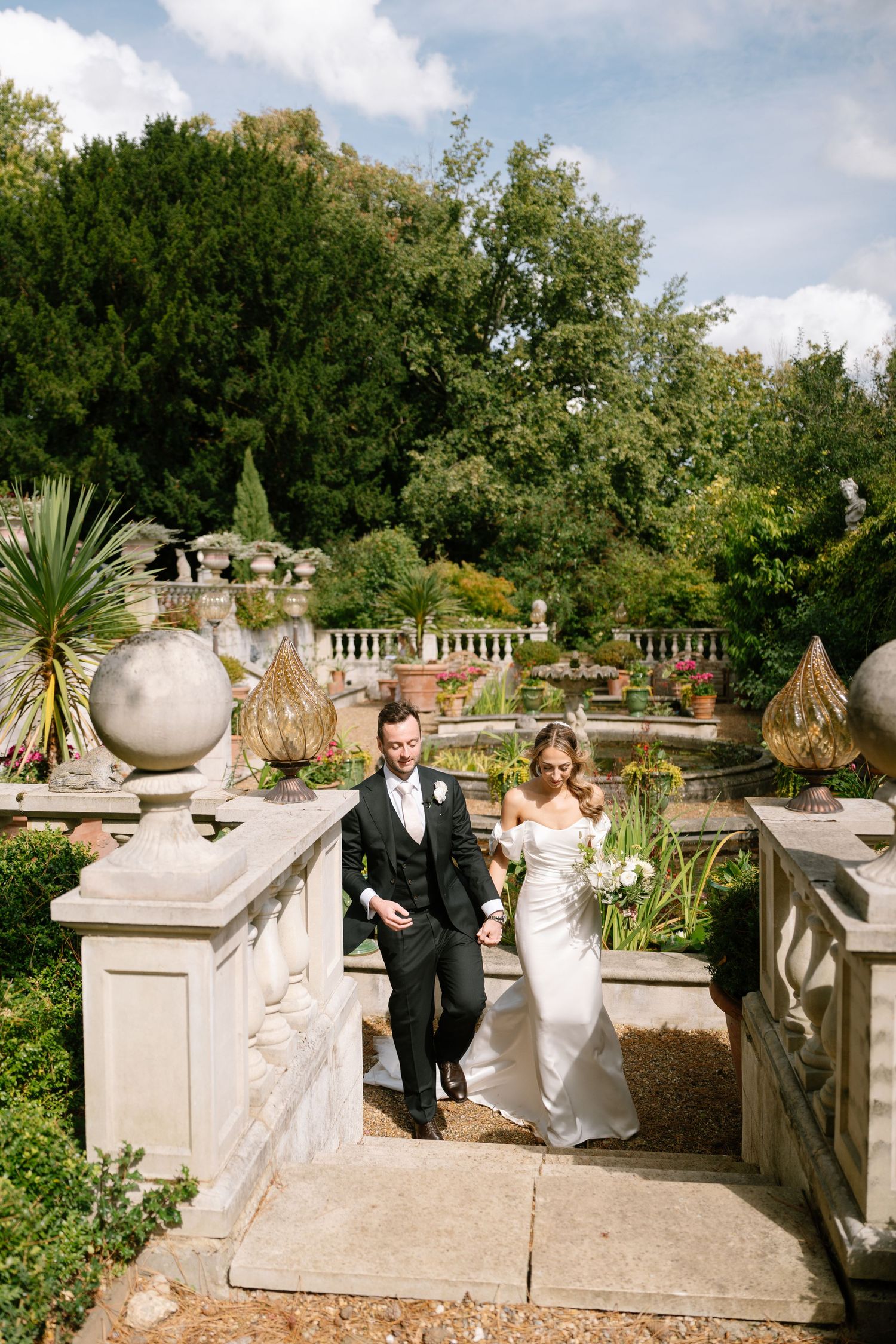 Wedding couple posing on stone terrace surrounded by potted plants and garden decor.