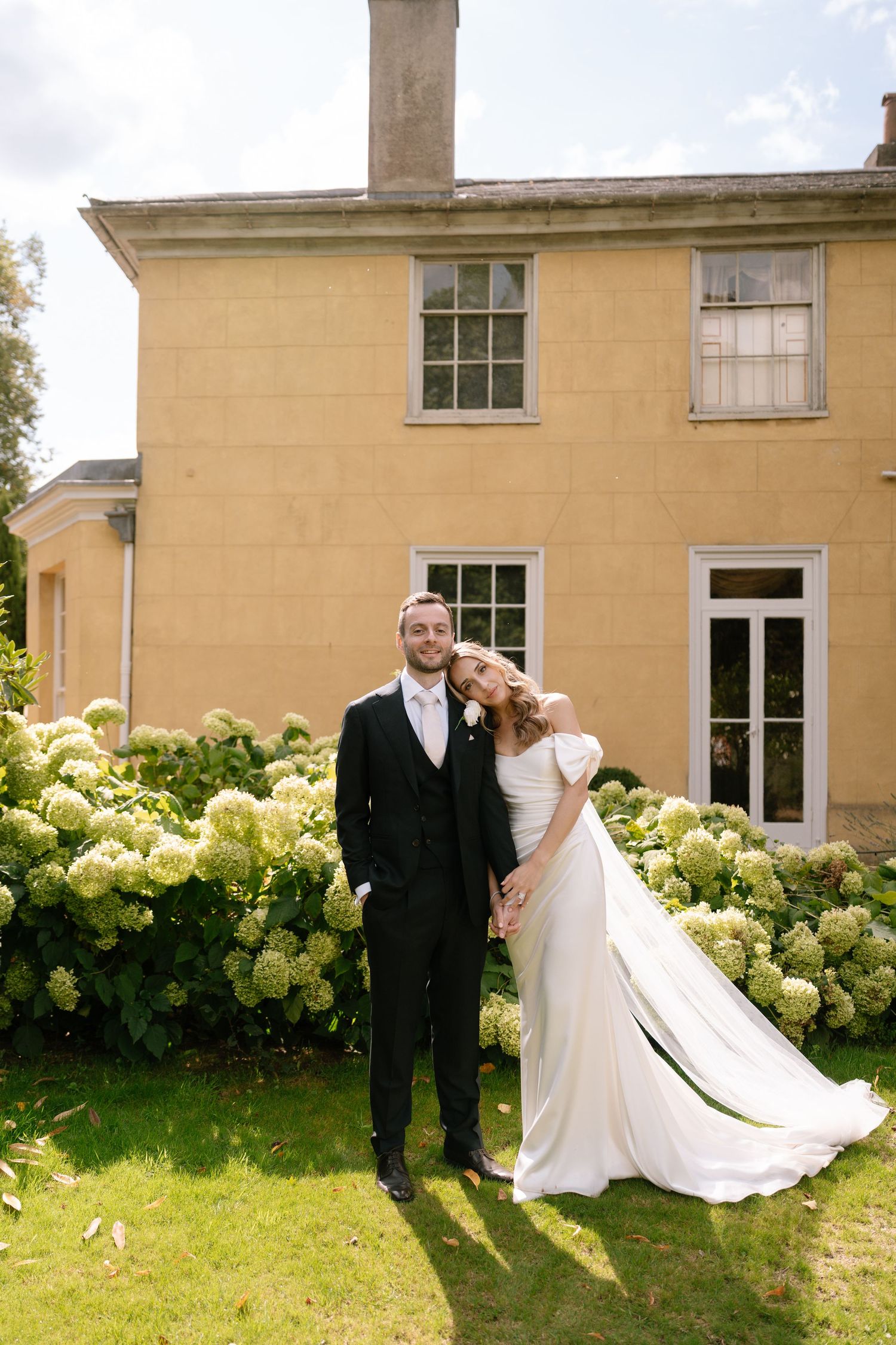 Wedding couple poses for romantic photos in front of a yellow building with hydrangea bushes.