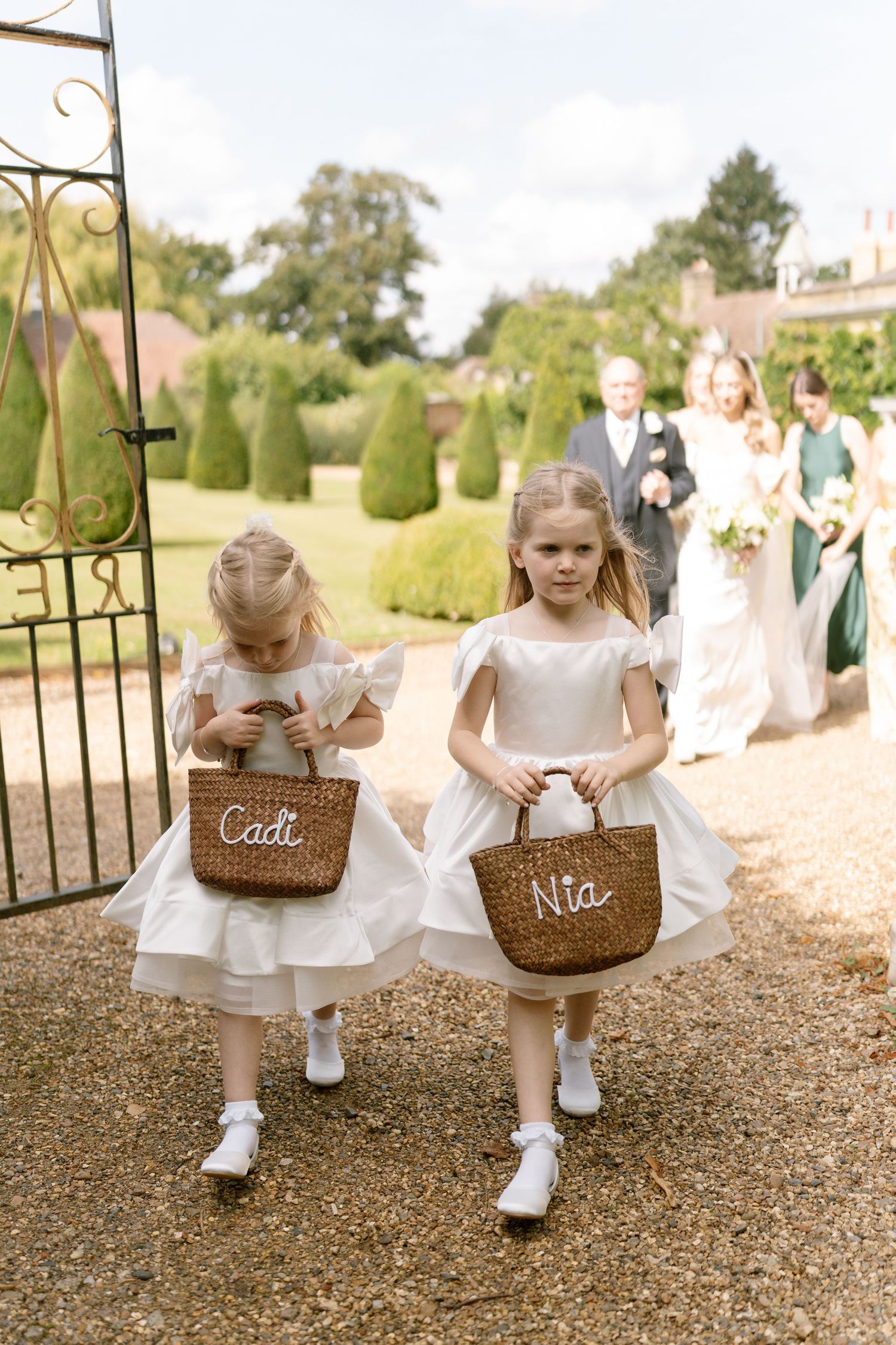 Flower girls in white dresses carrying wicker baskets through garden path.