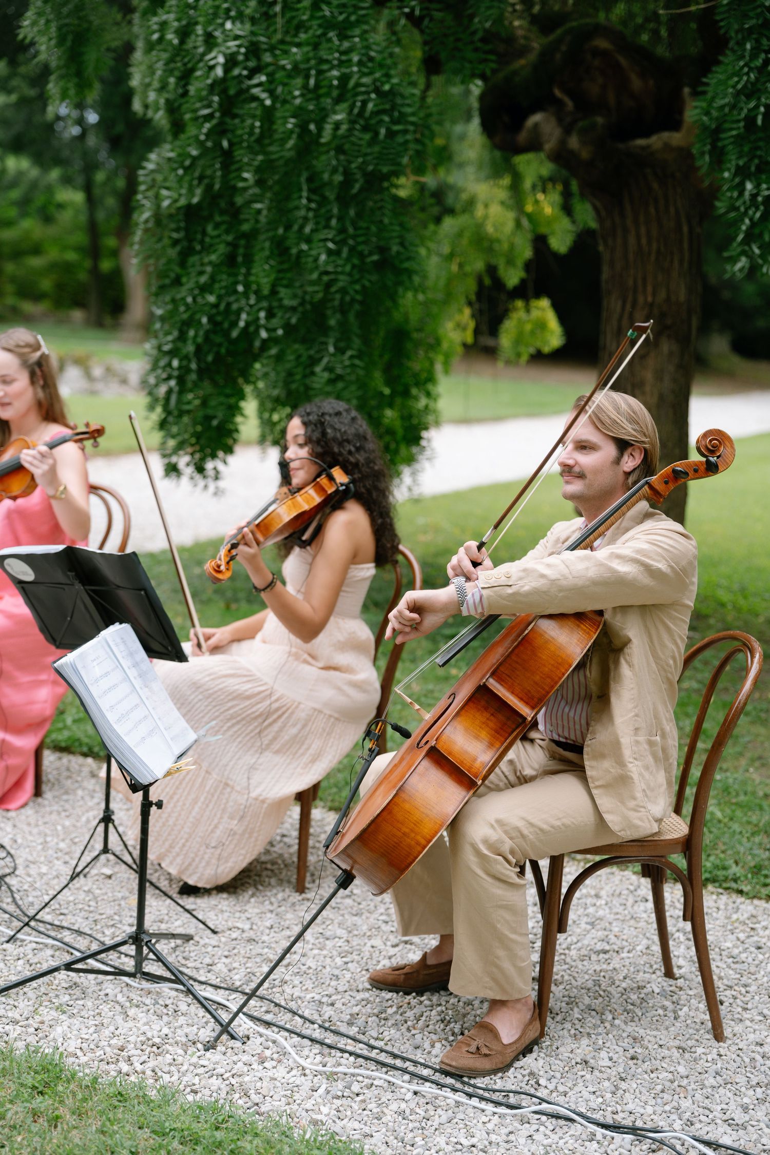 String musicians perform at an outdoor event while seated on vintage wooden chairs.