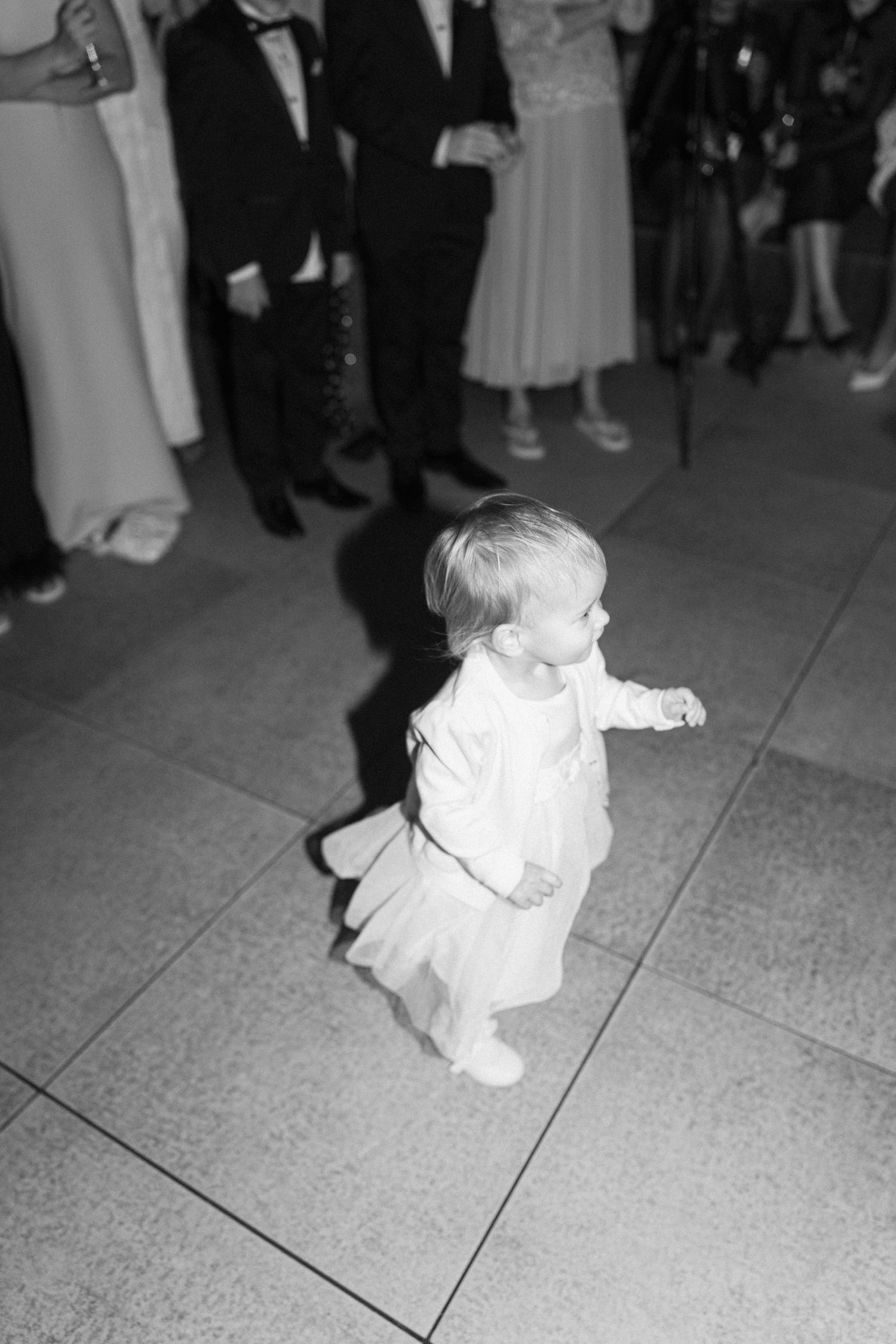 A toddler in white dress spins and dances on a tiled floor at an event.