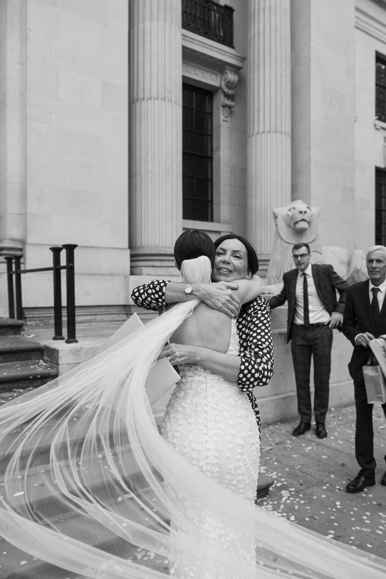 A bride in a polka dot dress twirls her long veil dramatically against a columned building.