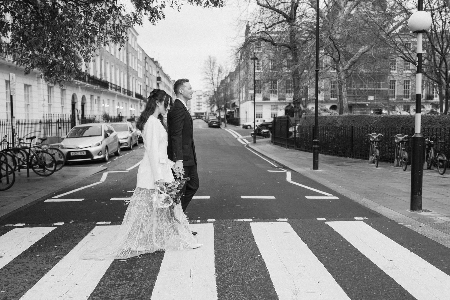 Black and white photo of a bride and groom crossing a zebra crossing on a London street.