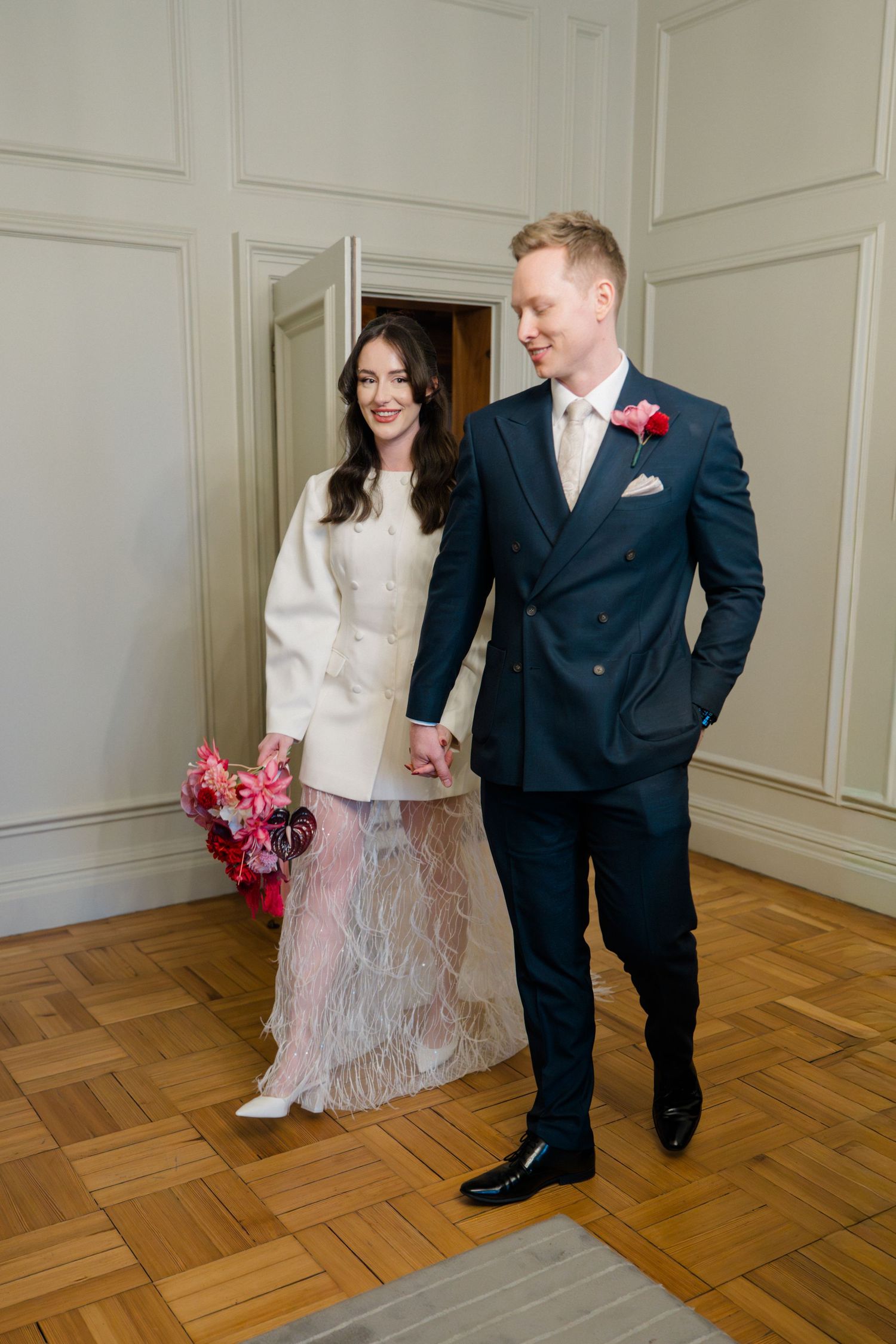 Smiling bride in white feather skirt holding pink bouquet walks hand in hand with groom in navy suit.
