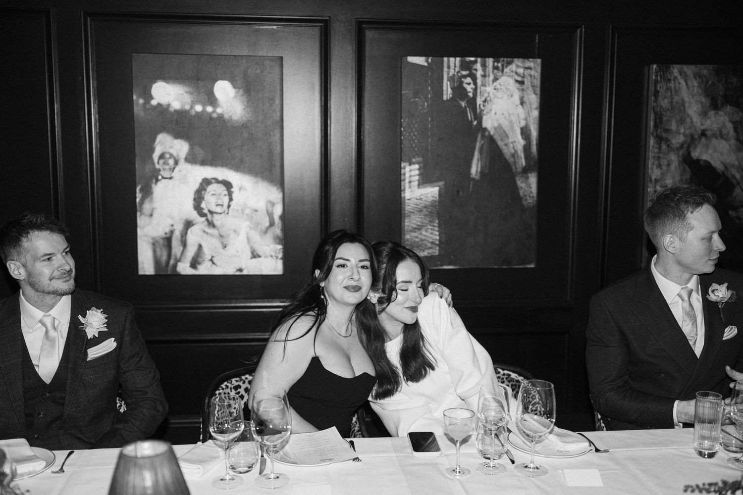 Wedding party seated at dinner table in black and white photo, bride in white and guest in black sharing a moment.