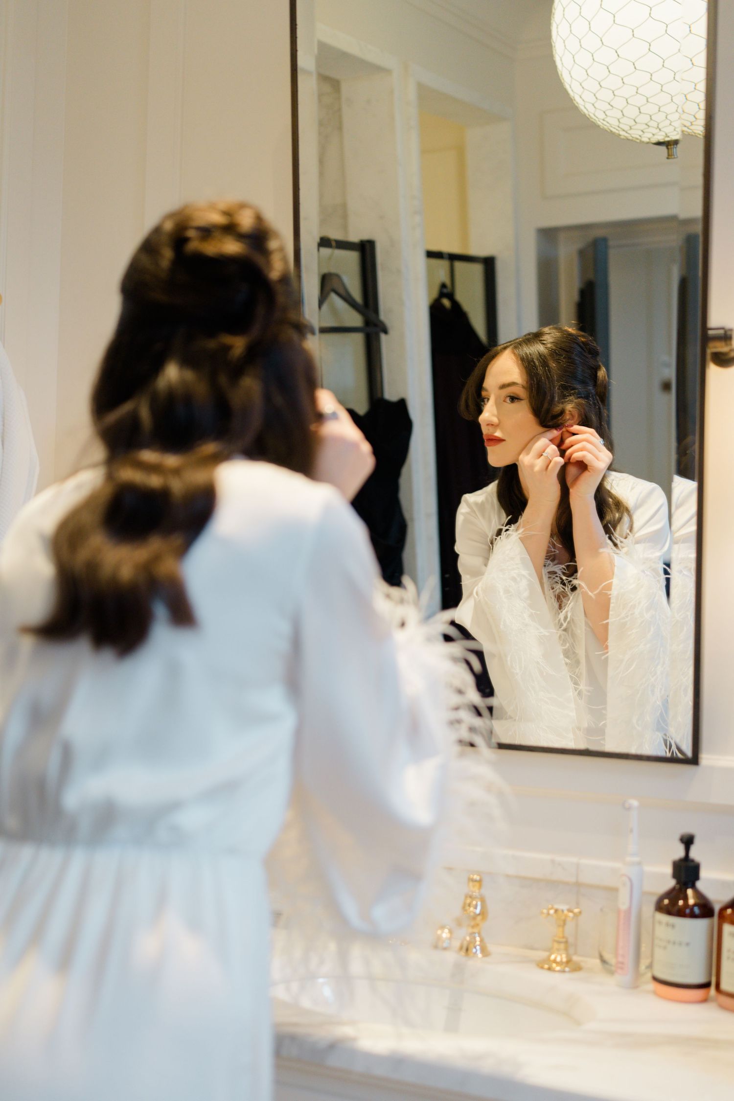 Bride in white lace robe putting on earrings while looking in bathroom mirror.