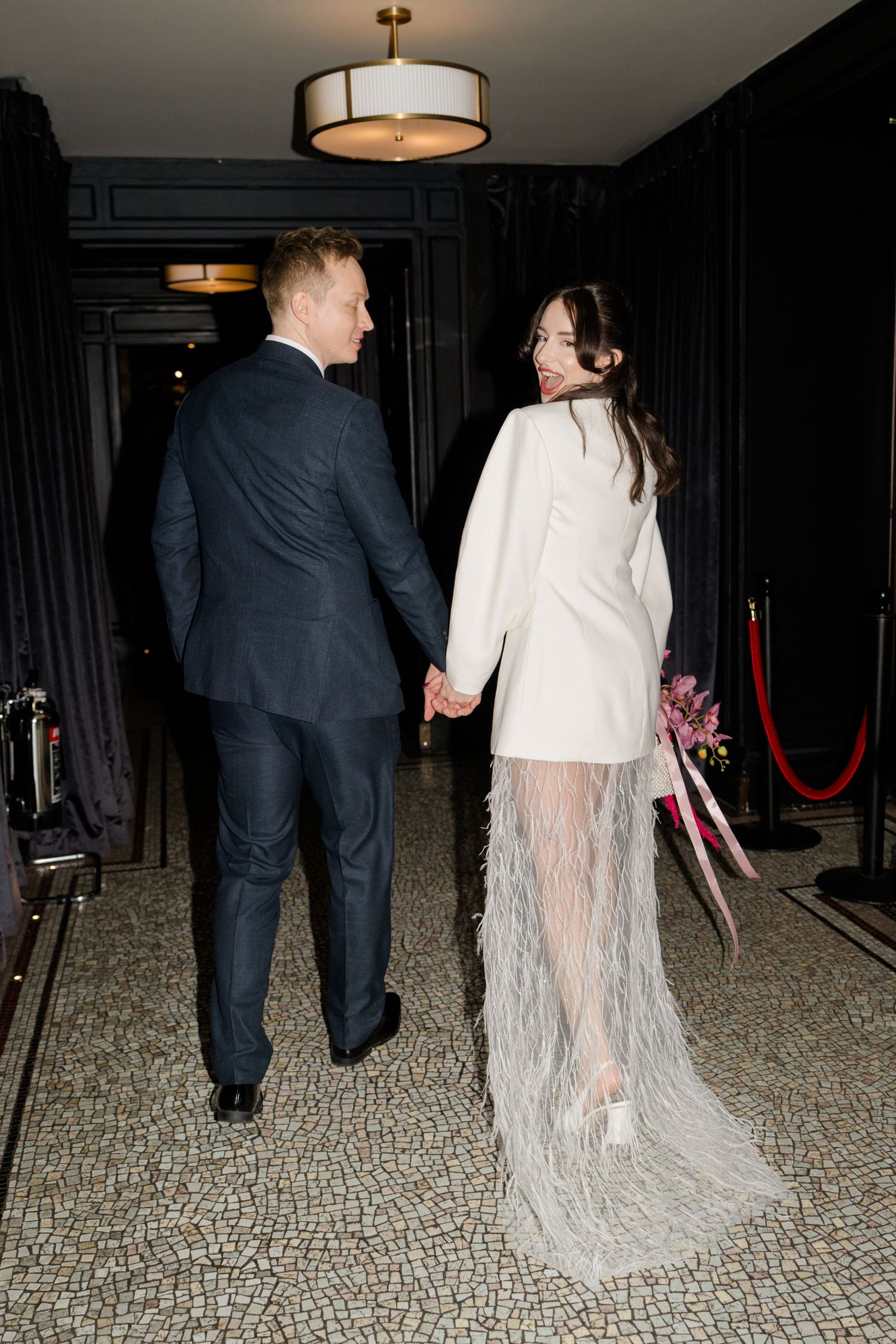 Bride in white feathered wedding dress holds hands with groom in navy suit walking down hotel corridor.