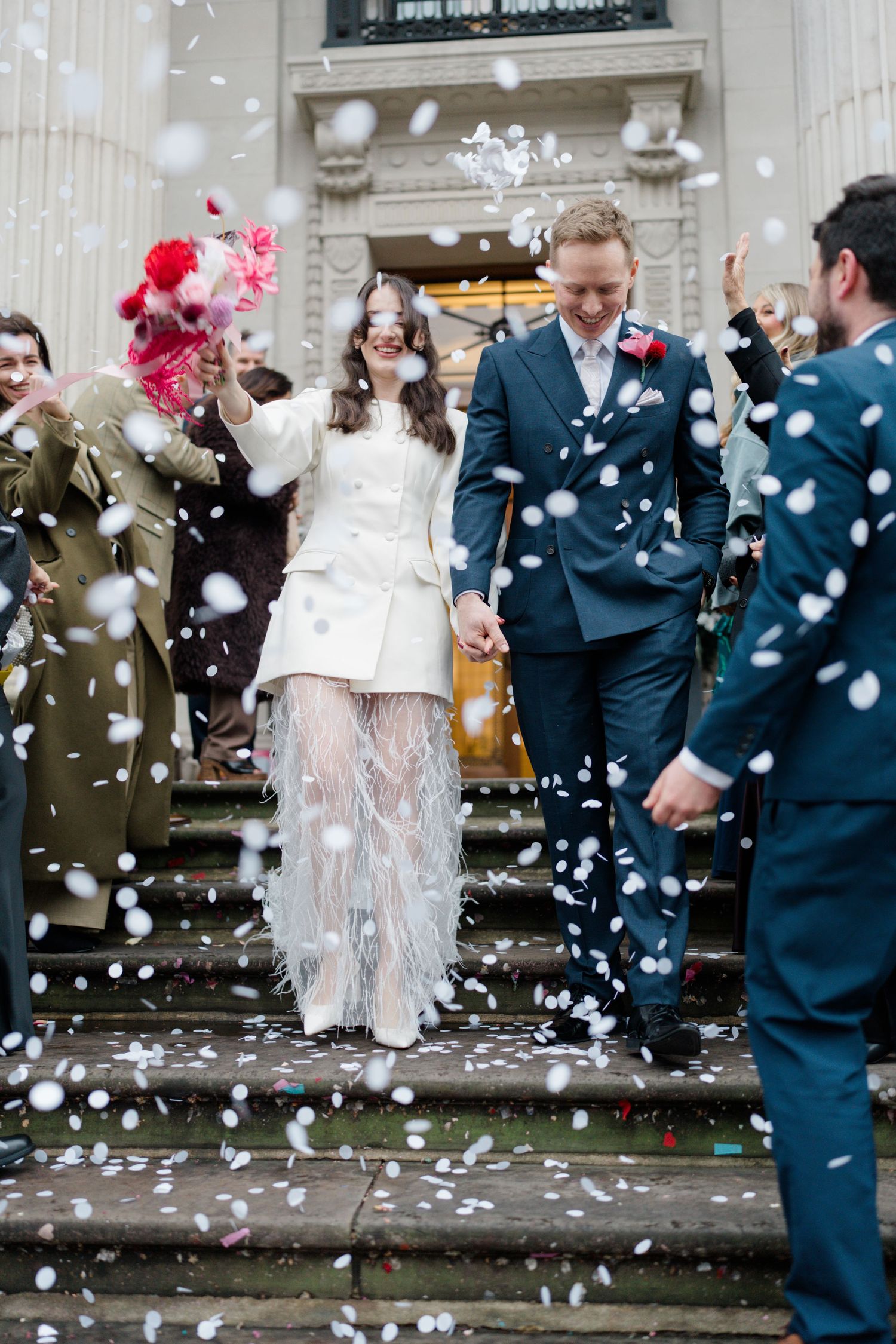 Bride and groom walk through confetti shower outside venue, bride wearing white feathered wedding outfit.