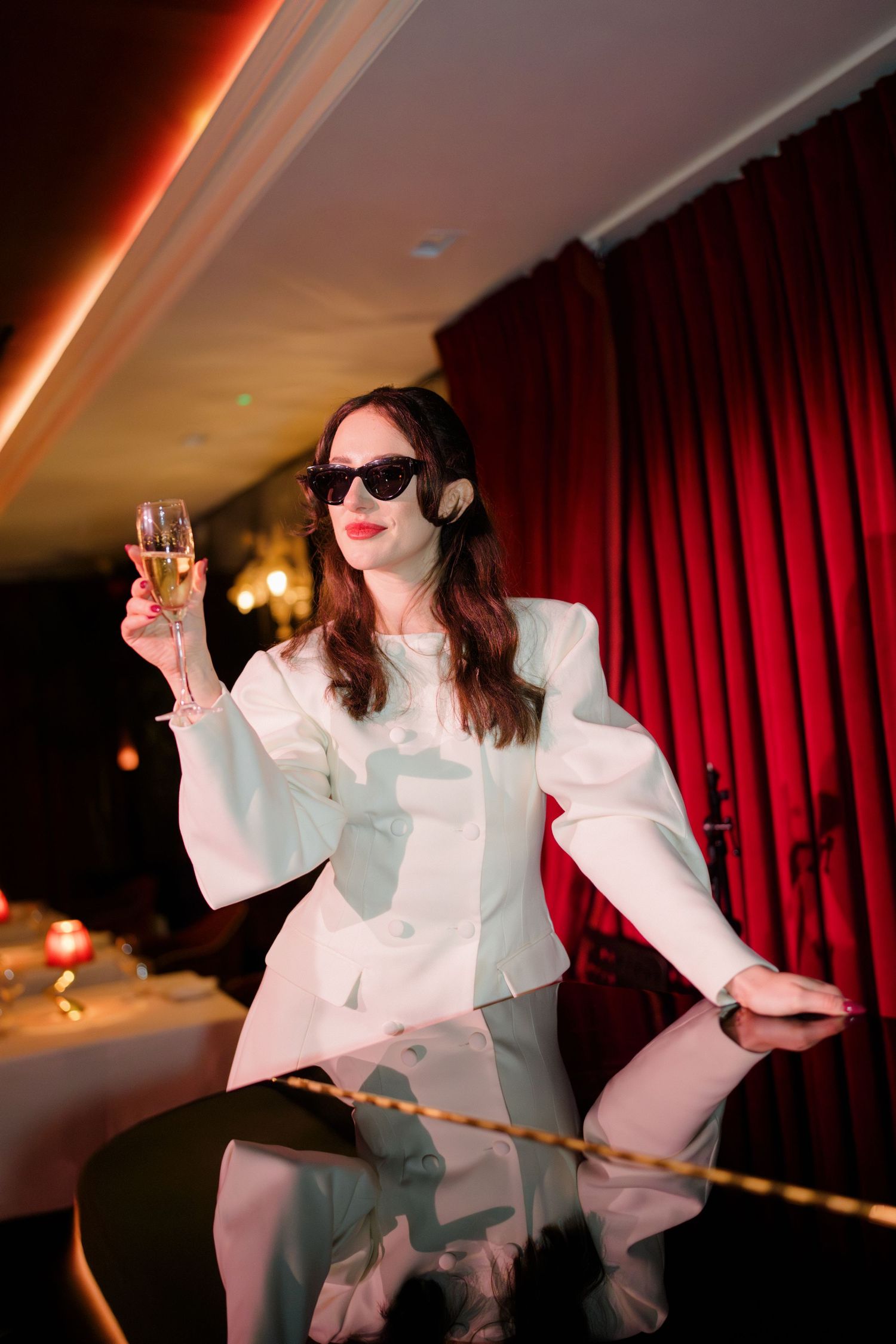 Bride in white structured dress wearing sunglasses raises champagne glass in front of red velvet curtains.