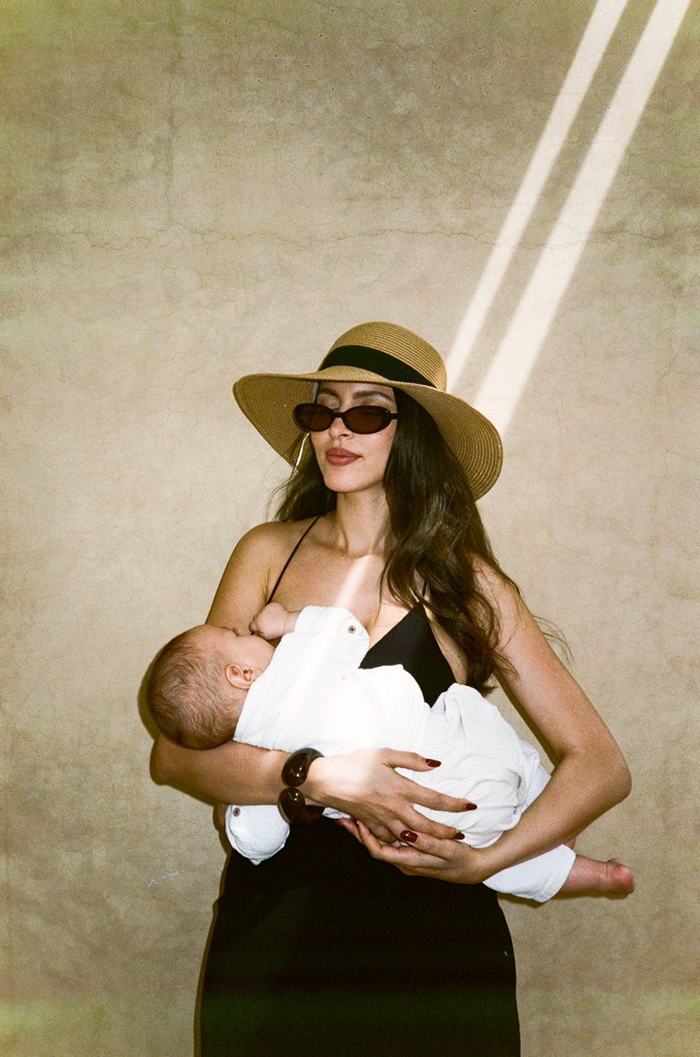 Stylish woman in sun hat and sunglasses breastfeeding newborn baby, wearing black dress outdoors.