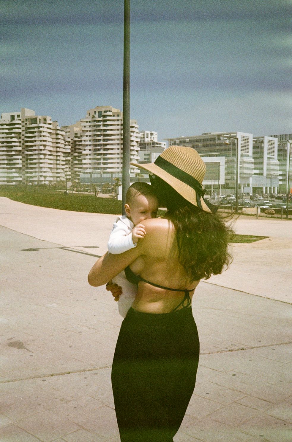 Woman in black backless dress and straw hat holding a baby outdoors near a coastal city promenade on a sunny day.