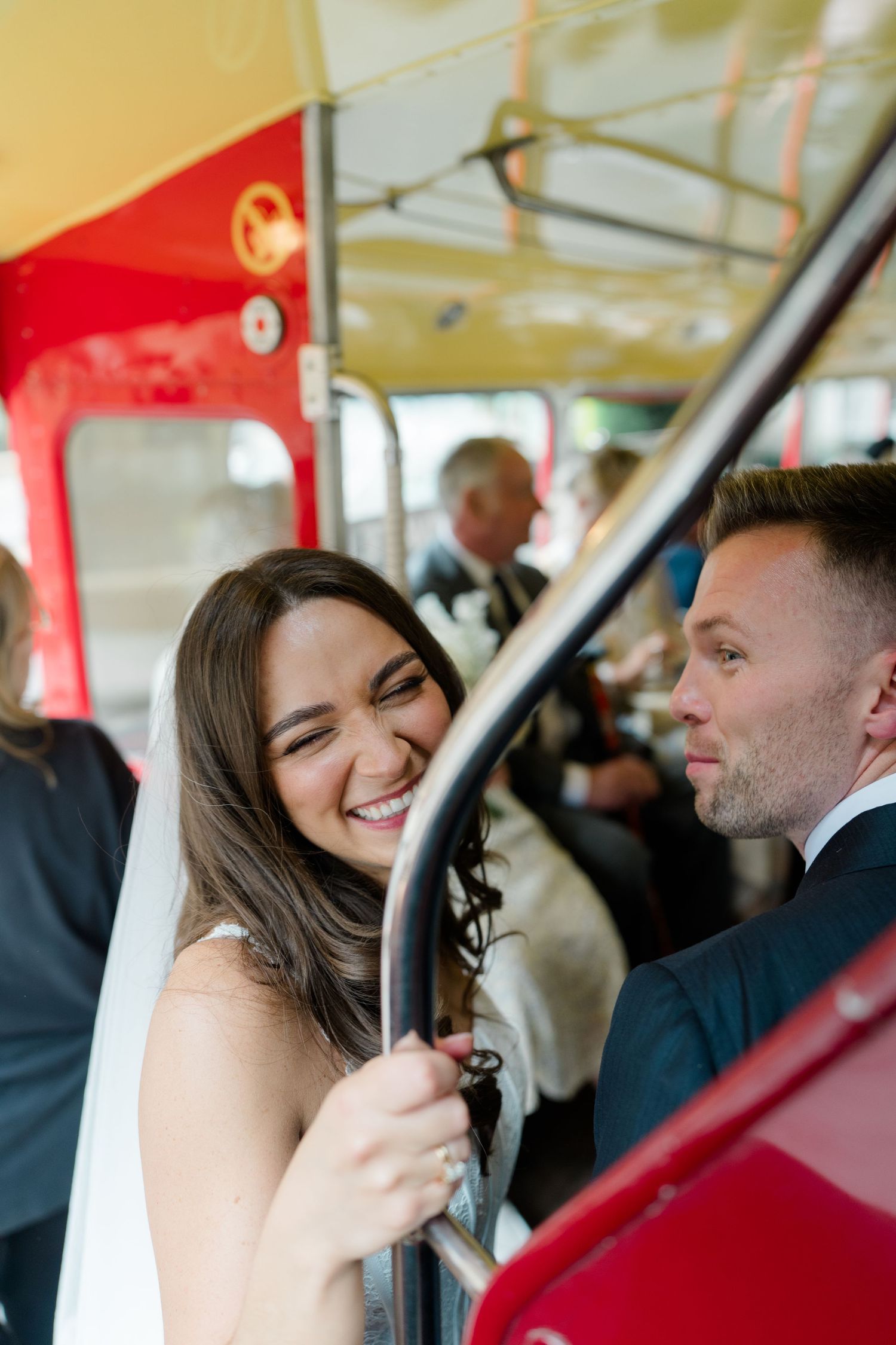 Smiling bride in veil laughs with groom inside vintage red double-decker bus at wedding celebration.