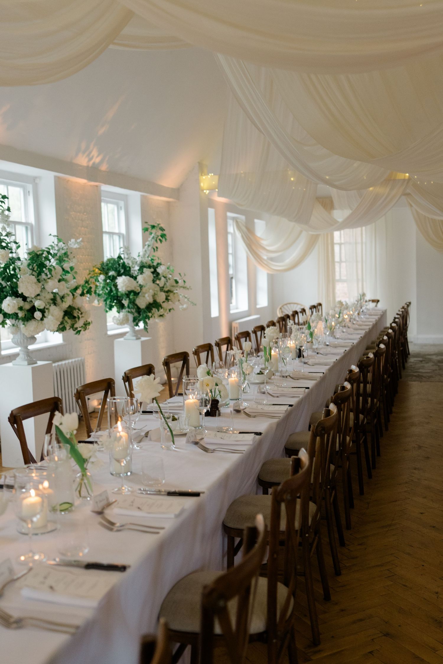 Elegant wedding reception hall with long white-draped tables, wooden chairs, tall floral centerpieces, and candles.