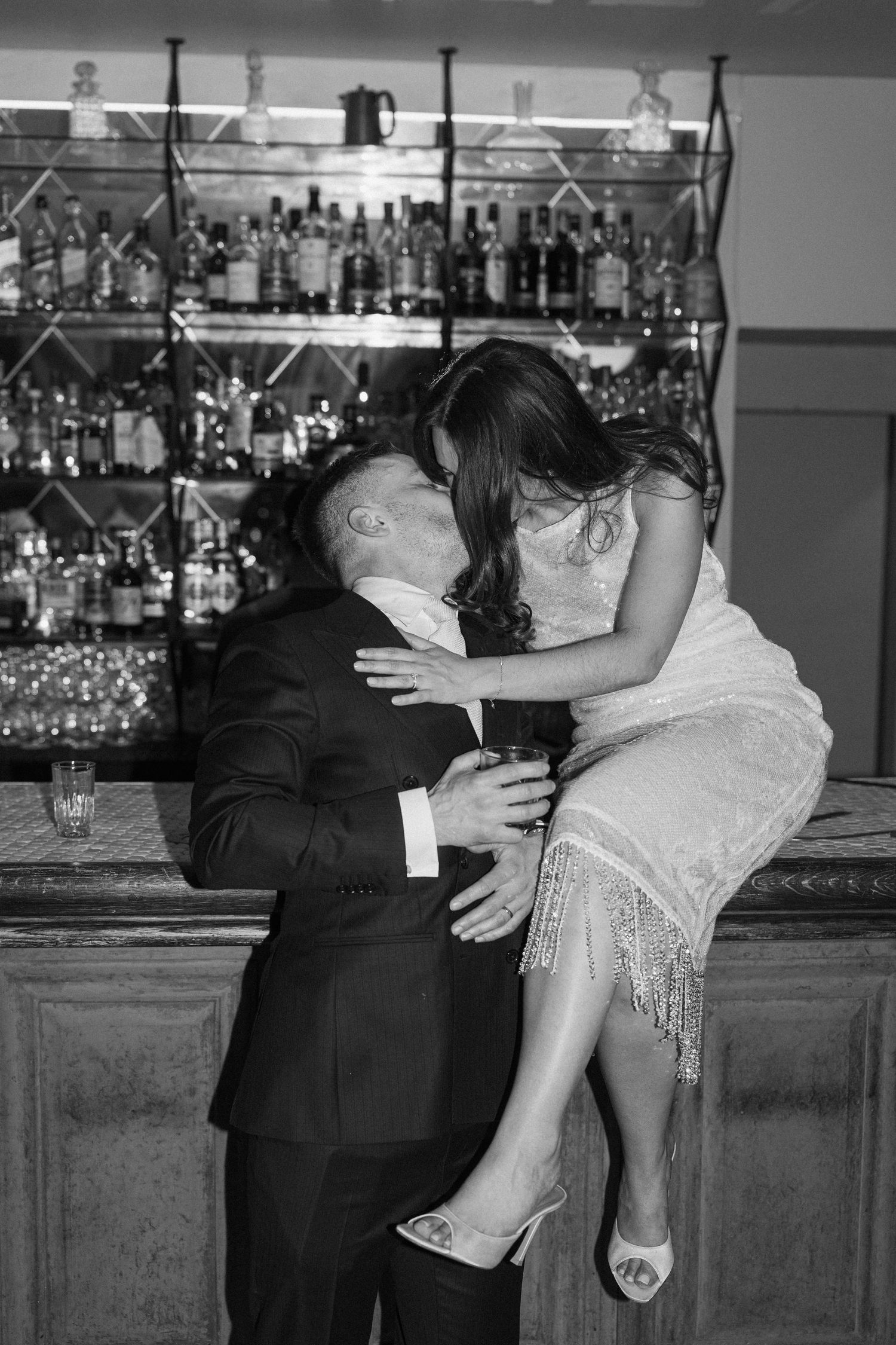 Black and white photo of groom kissing bride seated on bar counter at wedding reception venue.